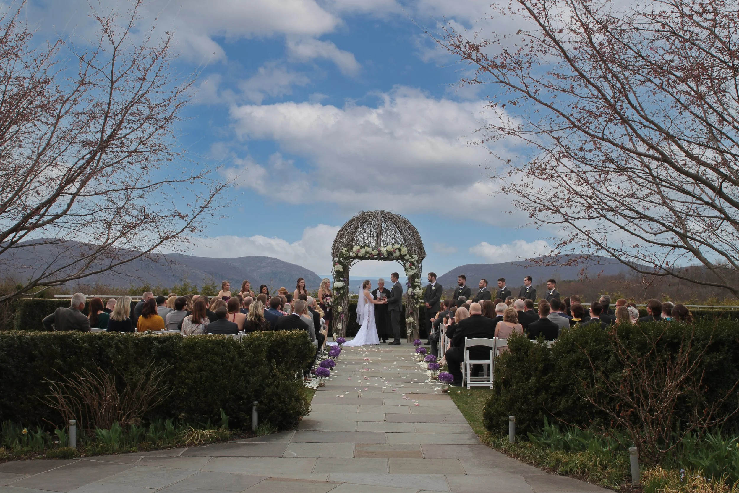 An outdoor wedding ceremony with a bride and groom standing under a wooden arch, surrounded by guests seated on white chairs, with mountains and a partly cloudy sky in the background at the Garrison.
