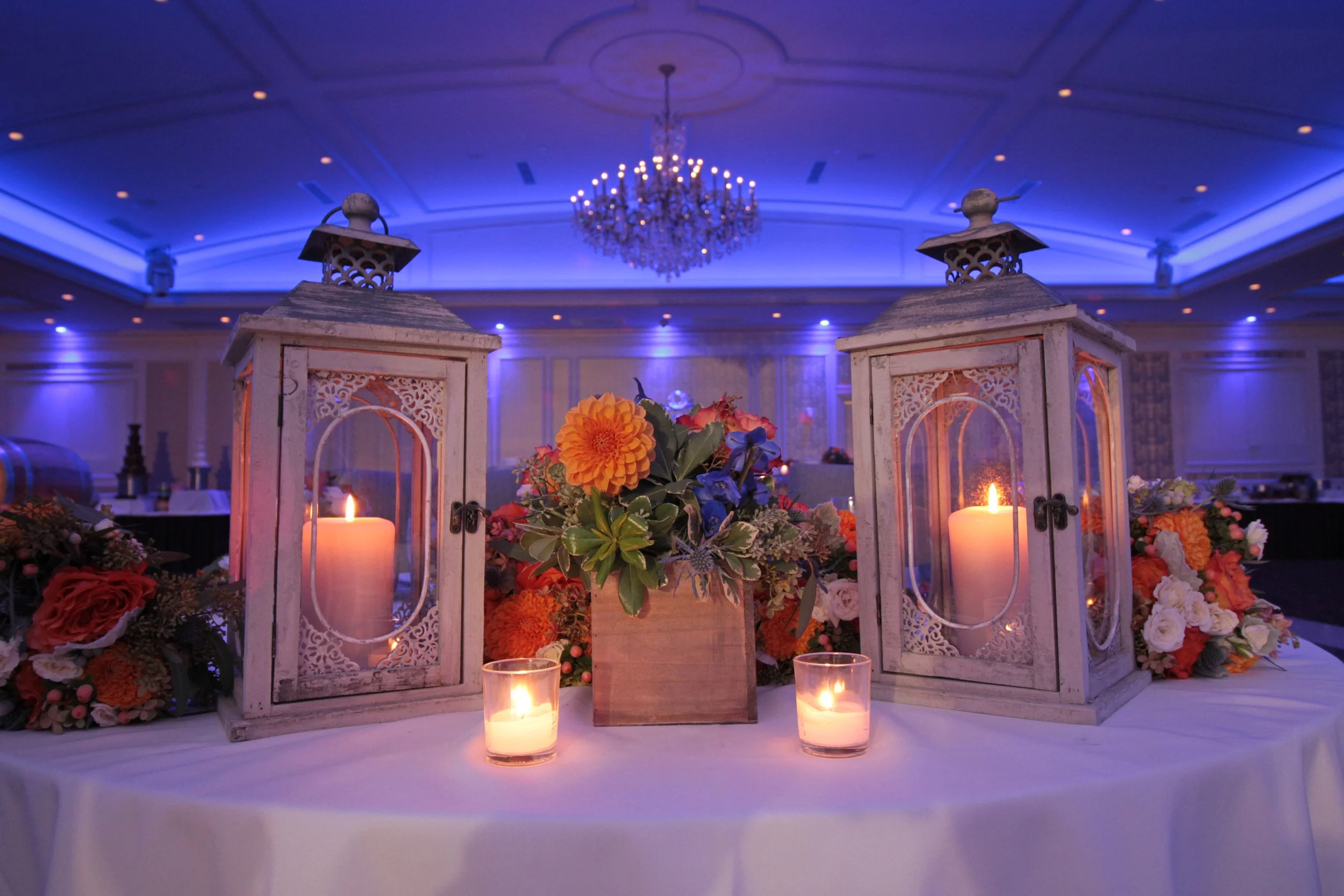 A decorated table with two white lanterns, three lit candles, and a floral arrangement in a wooden box, all set against a backdrop of an elegant ballroom with a chandelier and blue lighting.