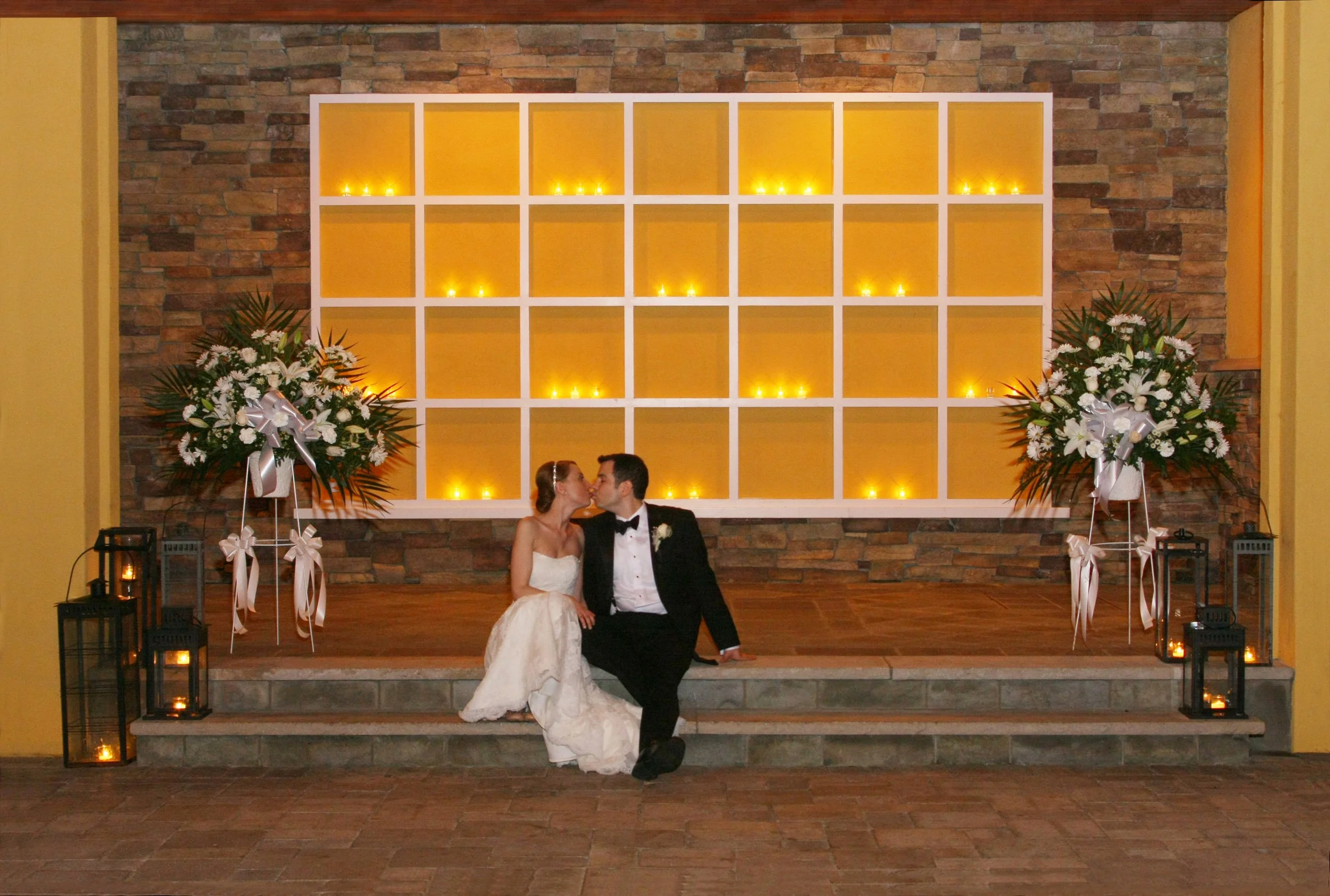 A bride and groom sit on steps in front of a backdrop made of a large white grid with yellow lights behind it. The bride is in a white wedding gown, and the groom is in a black tuxedo. Large floral arrangements with white flowers and greenery, decora