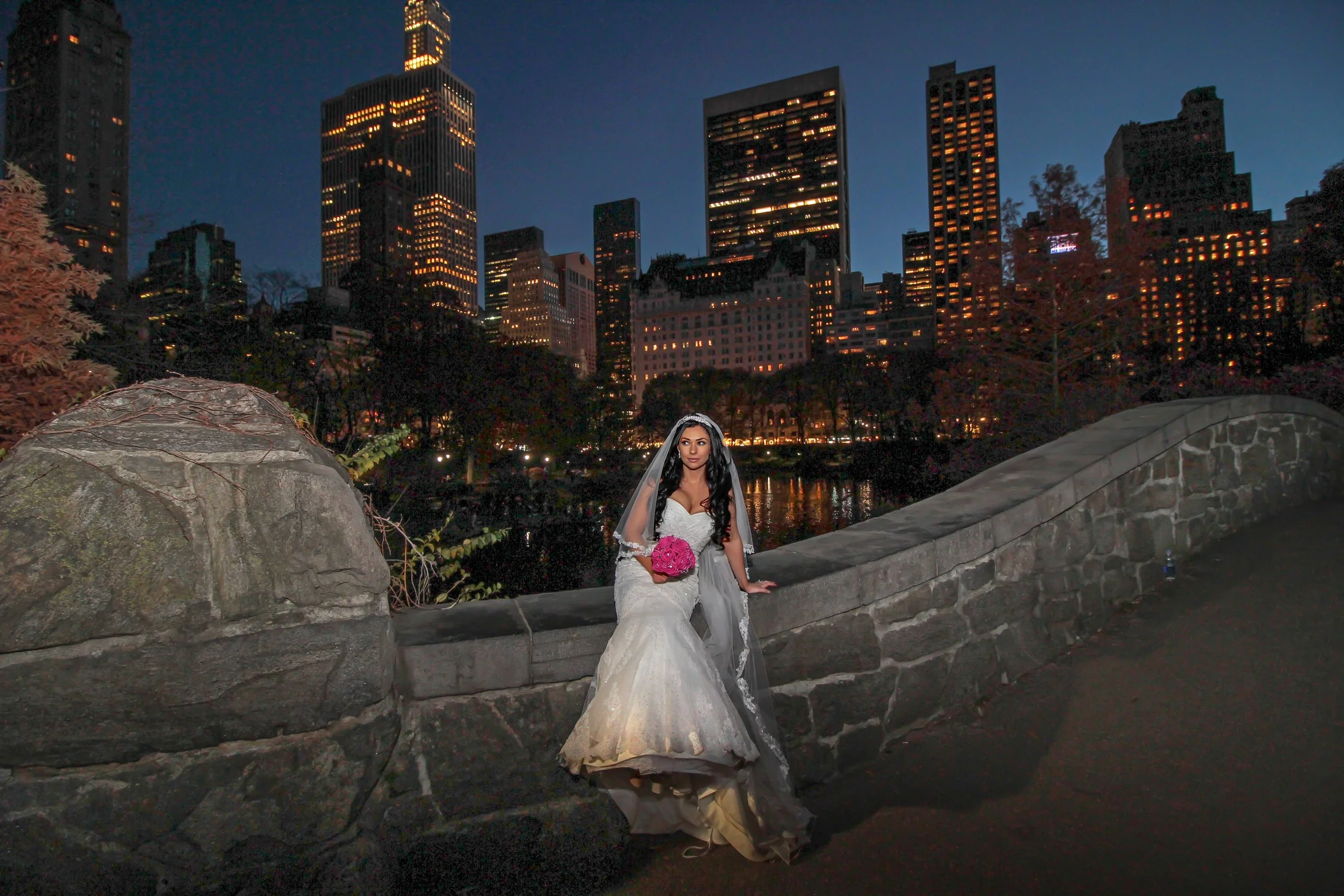 A woman in a wedding dress holding a pink bouquet stands on a stone bridge in an urban park at dusk, with city skyscrapers illuminated in the background.