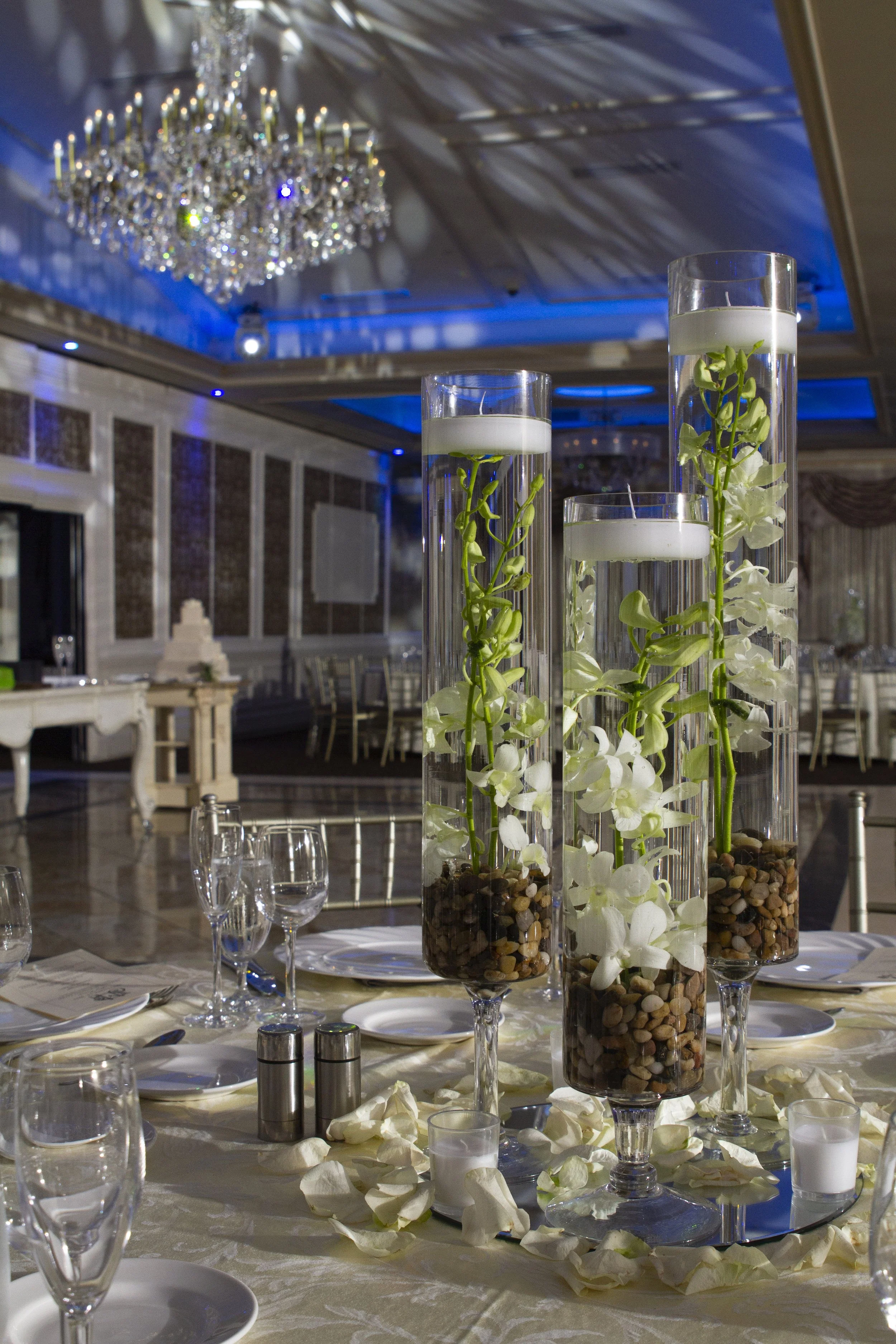 Elegant banquet table with tall glass vases filled with water, white orchids, and pebbles, surrounded by scattered white flower petals, set in a decorated event hall with a large chandelier.