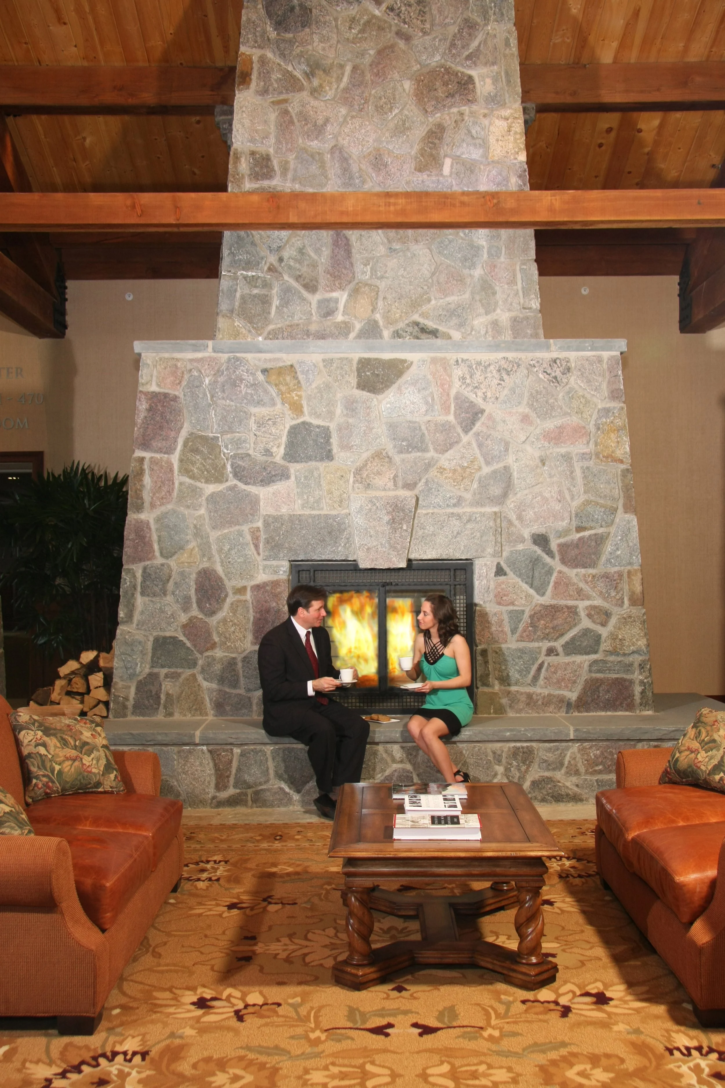 A man and a woman sit on a low ledge in front of a large stone fireplace with a fire burning inside. They are holding cups and appear to be having a conversation. The room has wooden ceilings, a patterned carpet, and couches with floral cushions.