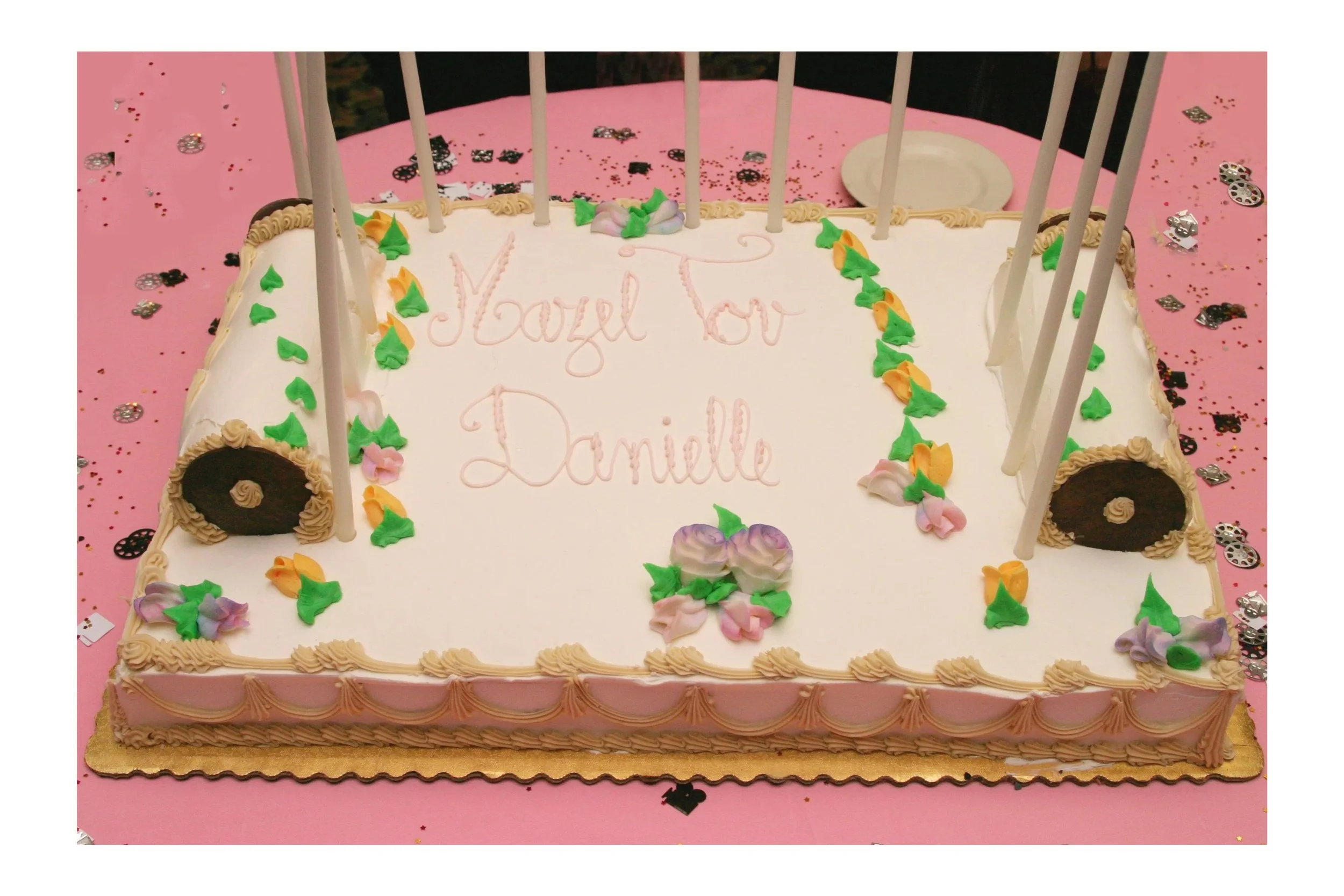 A Bat Mitzvah cake with white icing decorated with flowers and green leaves, with lit candles around the edges. Pink cursive writing on the cake says, "Lovely To Danielle." The cake is on a pink tablecloth with scattered silver and black confetti.