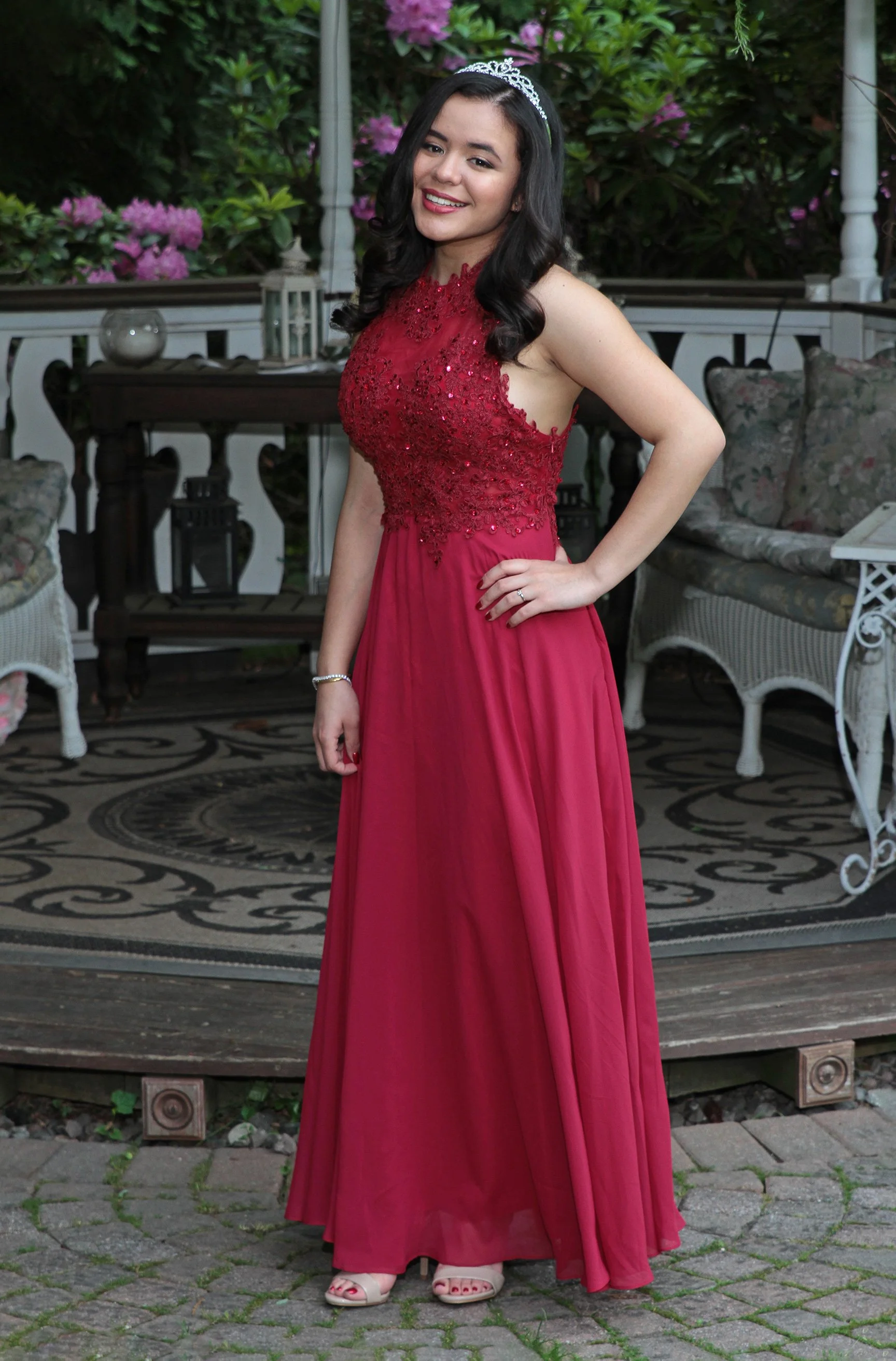 A young woman in a red formal gown with lace and beading details, wearing a tiara, standing outdoors on a stone patio with flowers and garden furniture behind her, smiling at the camera.
