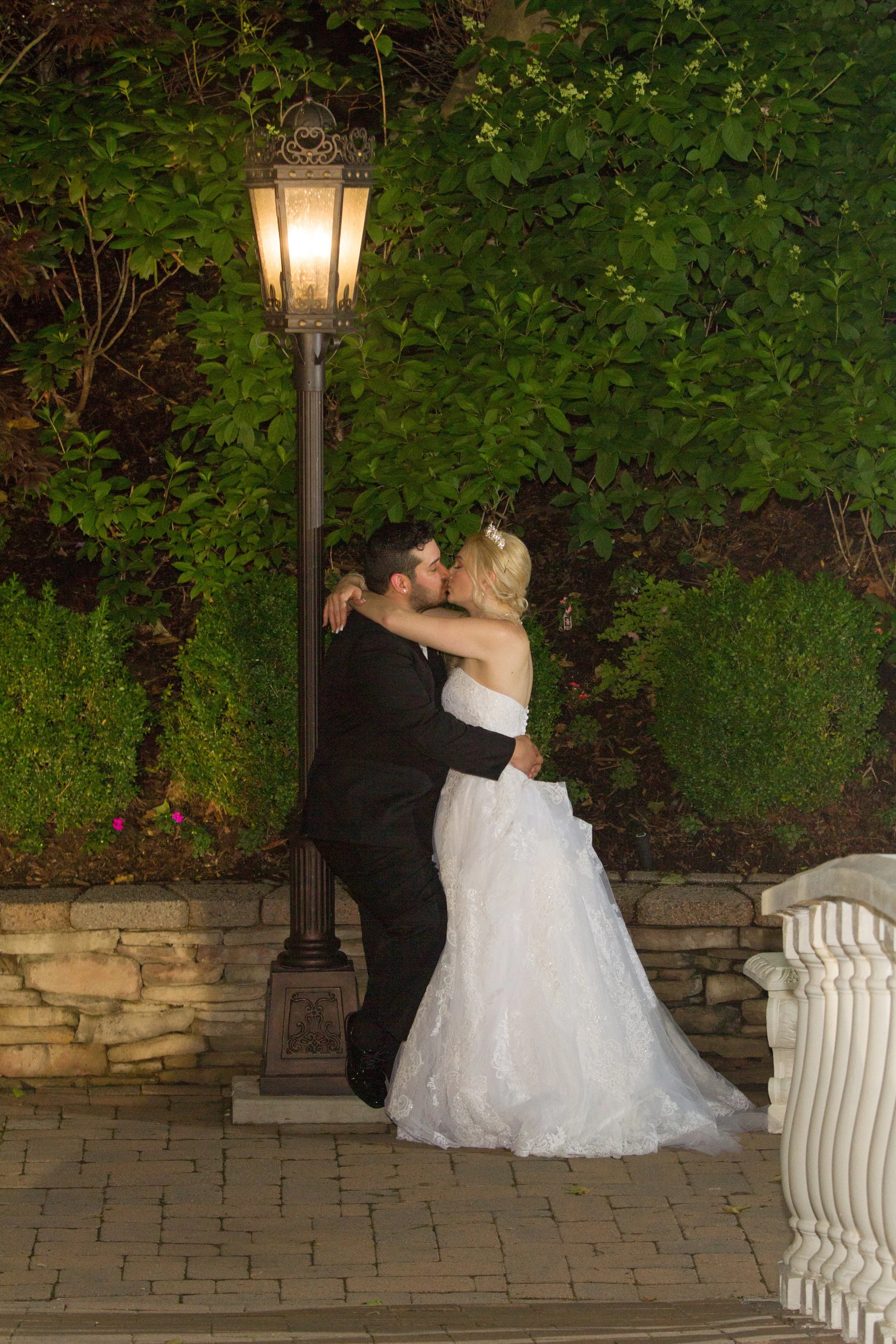 A bride and groom kissing outdoors at night, standing next to a lamppost, with greenery behind them.