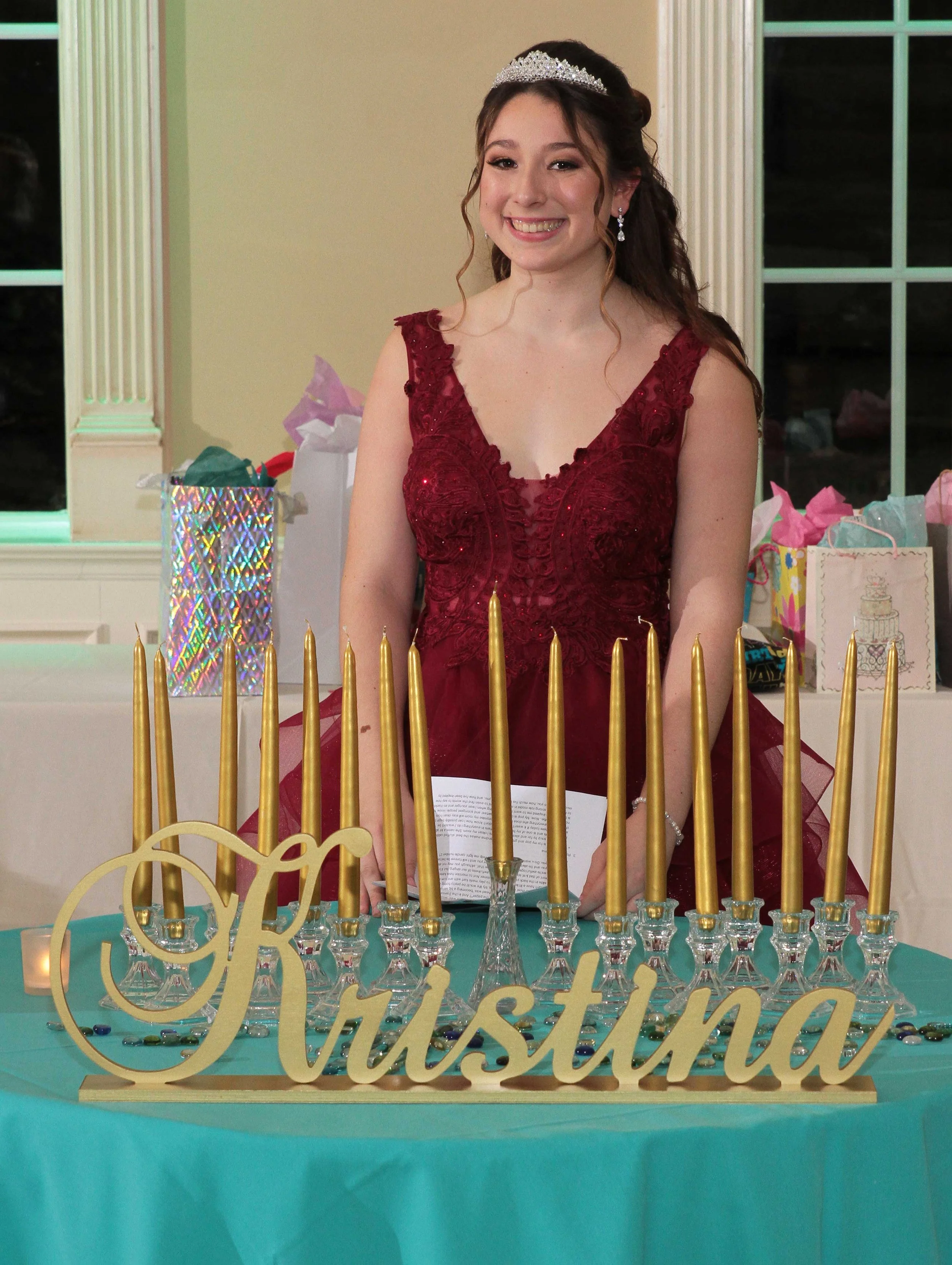 A young sweet sixteen woman in a red dress and tiara standing behind a table with a large gold "Kristina" sign, golden candles, and birthday presents in the background.