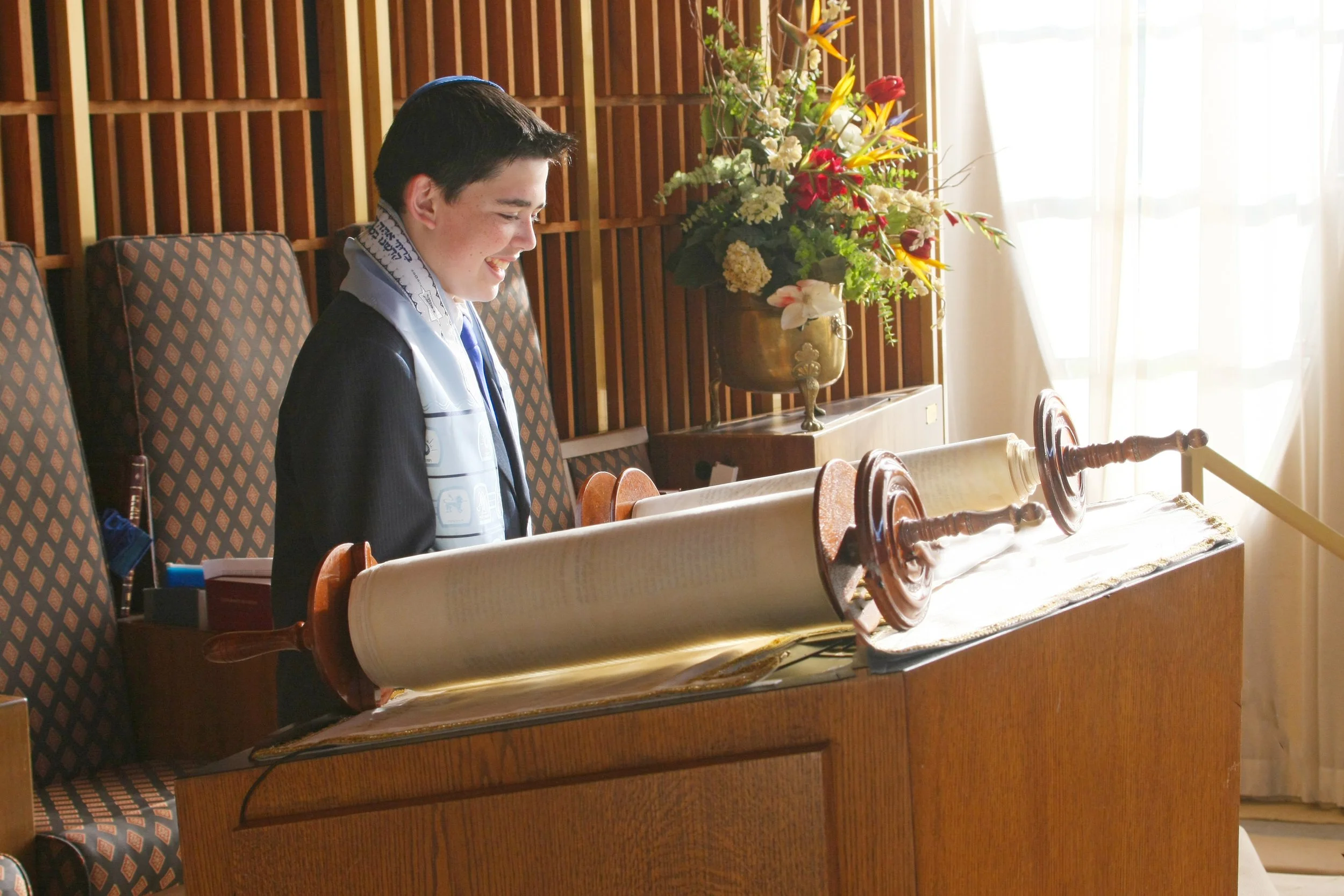 A young Bar Mitzvah boy wearing a kippah and a tallit standing at a Torah scroll in a synagogue, smiling.