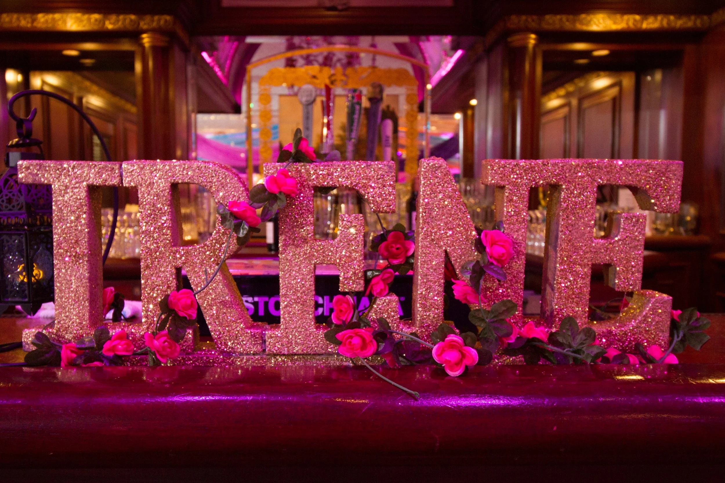 Decorative sweet sixteen sign spelling 'IRENE' in pink glitter with pink flowers, set in a dimly lit, elegant event space with pink and purple lighting.