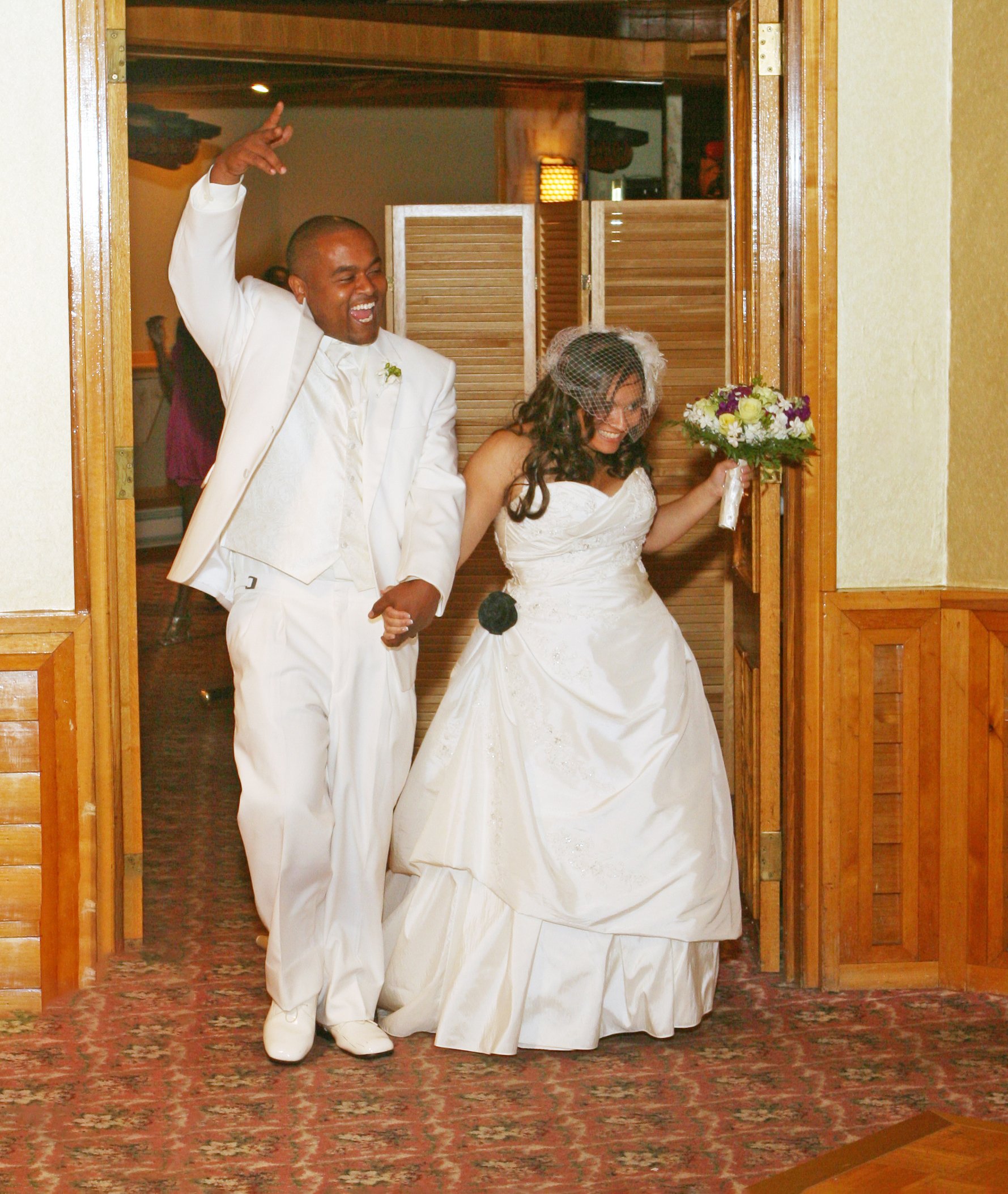 A bride in a white wedding gown holding a bouquet and a groom in a white suit entering a room, smiling and celebrating at the Views at Mount Fuji.