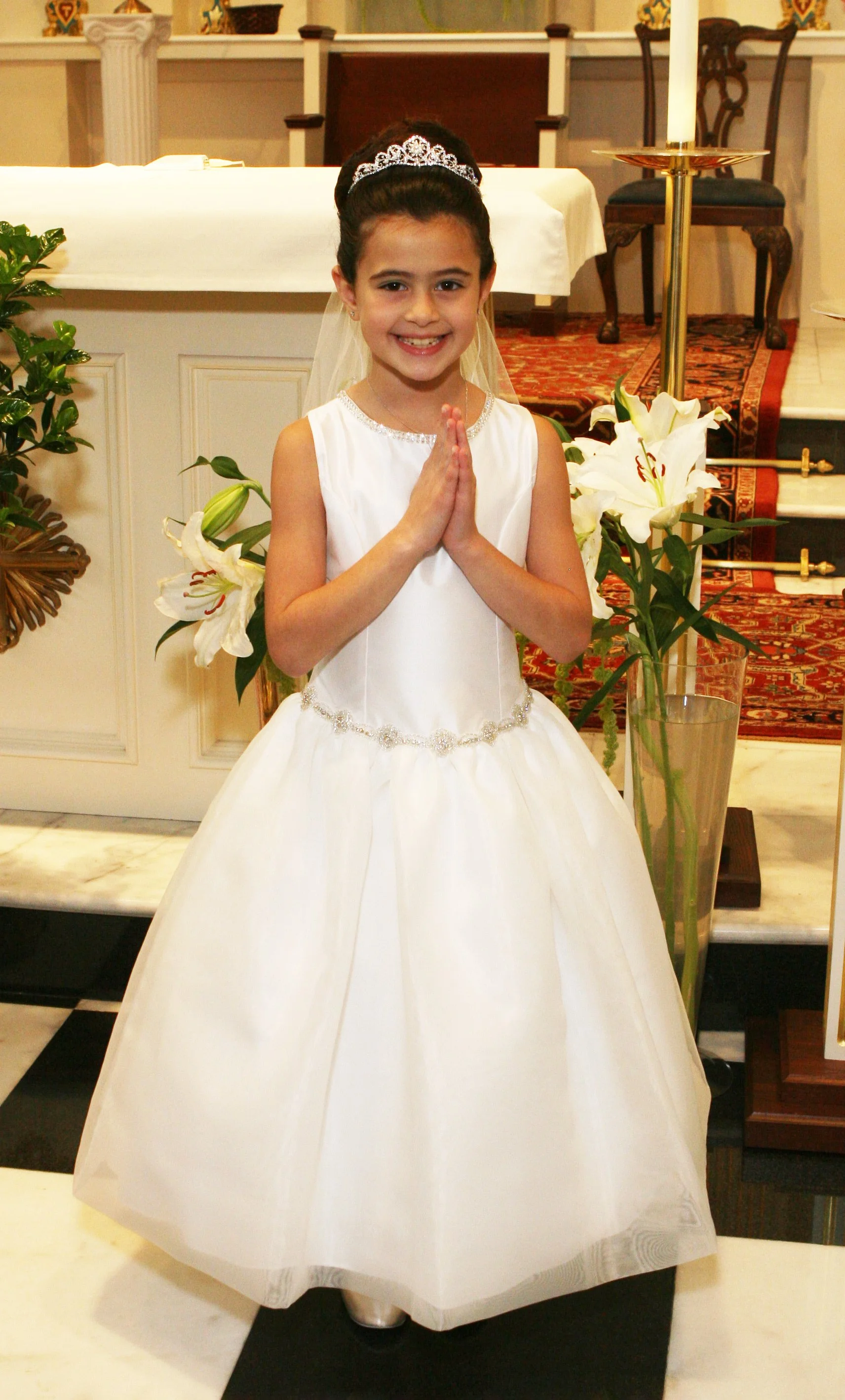 A young first communion girl in a white dress with a tiara and veil, standing on a black mat in front of St. Gabriel the Archangel's Roman Catholic church altar, smiling with hands clasped.