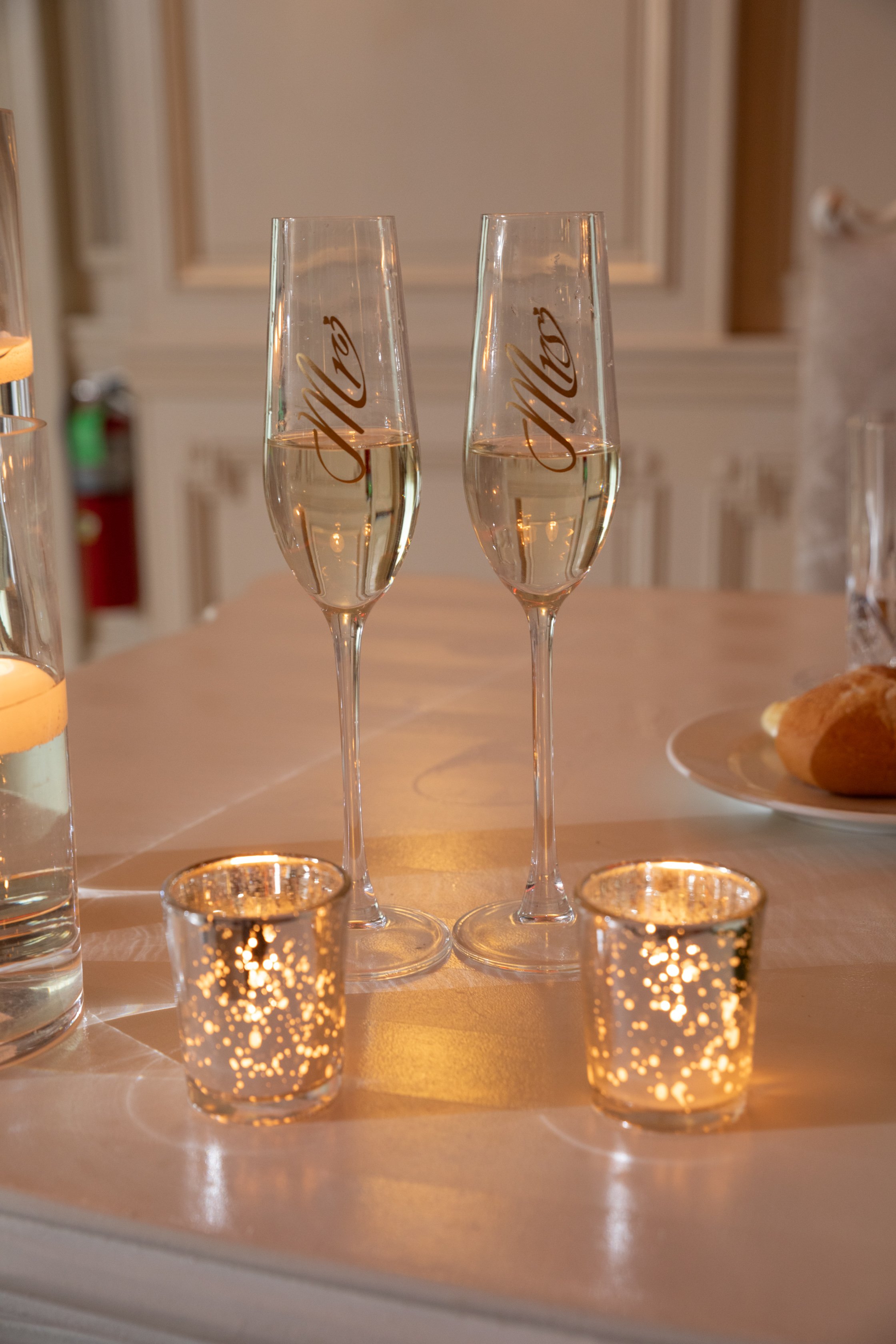 Two champagne flutes filled with champagne, with gold script writing, Mr. and Mrs., placed on a table with two small lit candle holders, surrounded by other drinking glasses and a plate of bread rolls in the background.