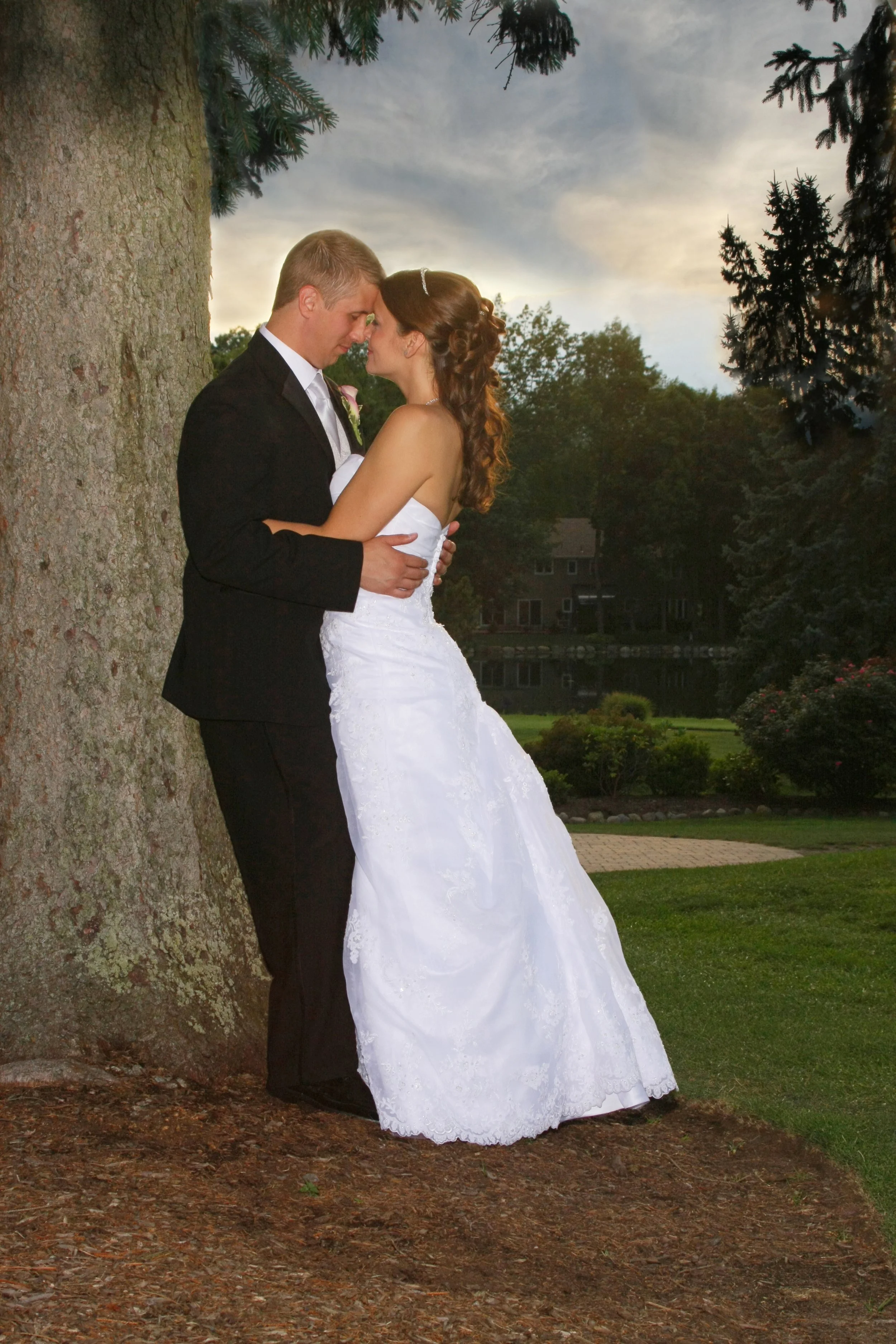 A bride and groom embrace next to a large tree outdoors at sunset, with water and trees in the background.