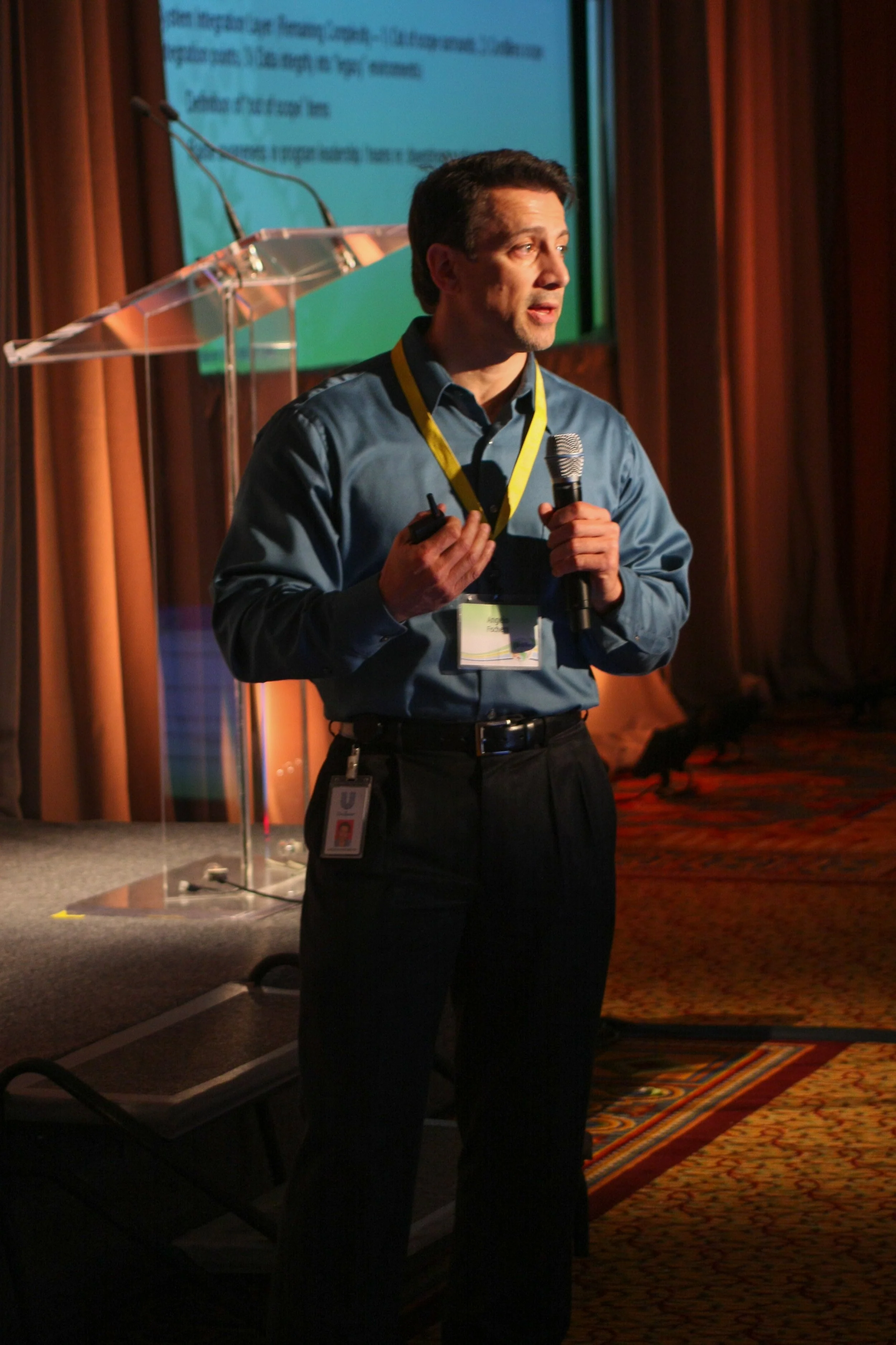 A man in a blue shirt holding a microphone and a remote control, standing on a stage with a presentation screen behind him. The room has wooden paneling and a patterned carpet.