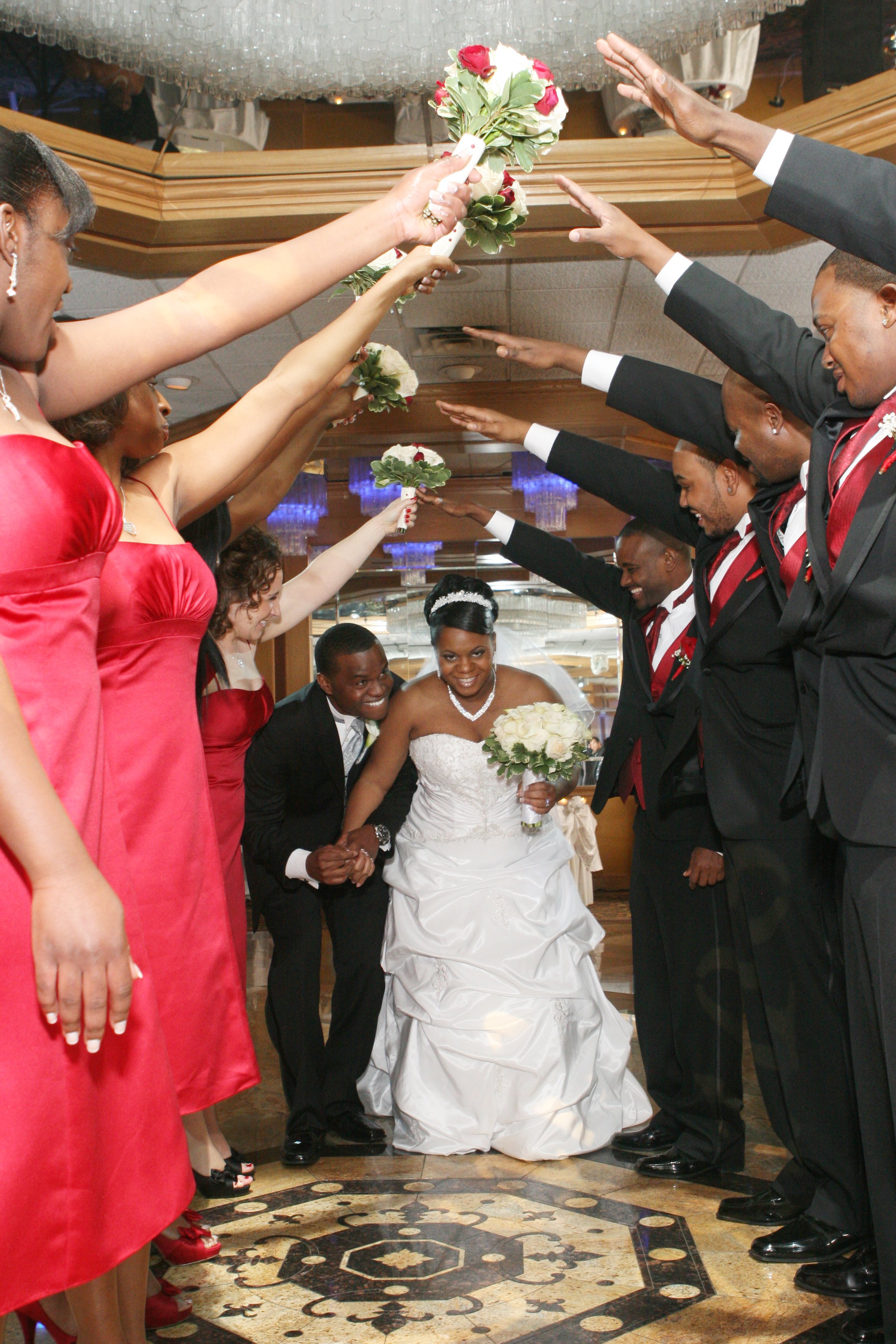 Happy bride in a strapless white wedding gown holding a bouquet, surrounded by bridesmaids in red dresses and groomsmen in black suits with red accents, forming an arch with their arms, on a decorative floor in a wedding reception hall.