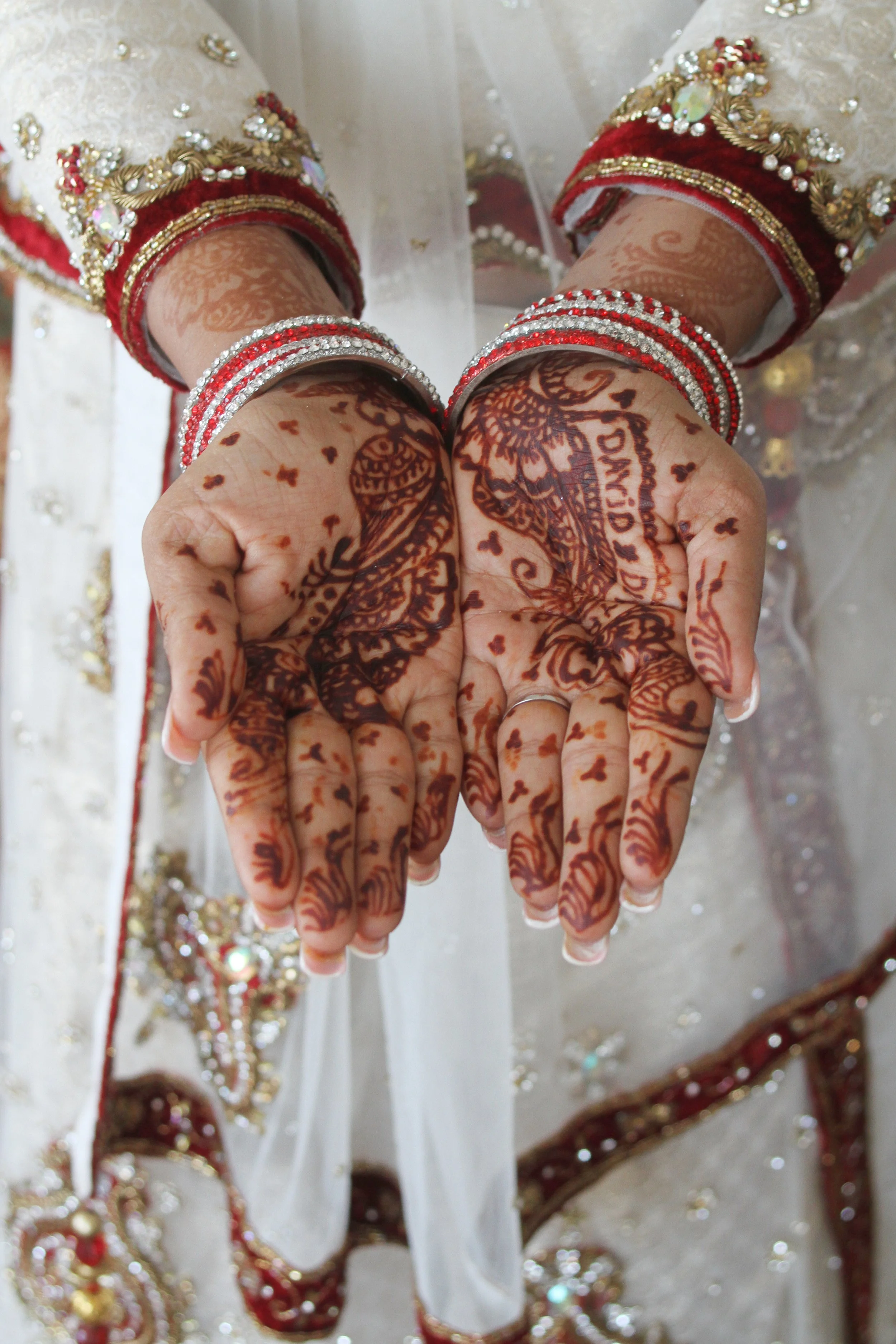 Close-up of hands with intricate henna designs, wearing silver and red bangles, and traditional South Asian bridal outfit with gold embroidery and beadwork.