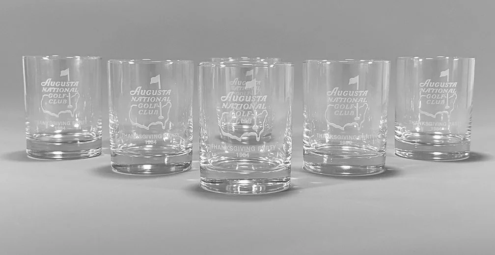 Six clear glass tumblers with the Augusta National Golf Club logo and text, arranged in a row on a gray surface.
