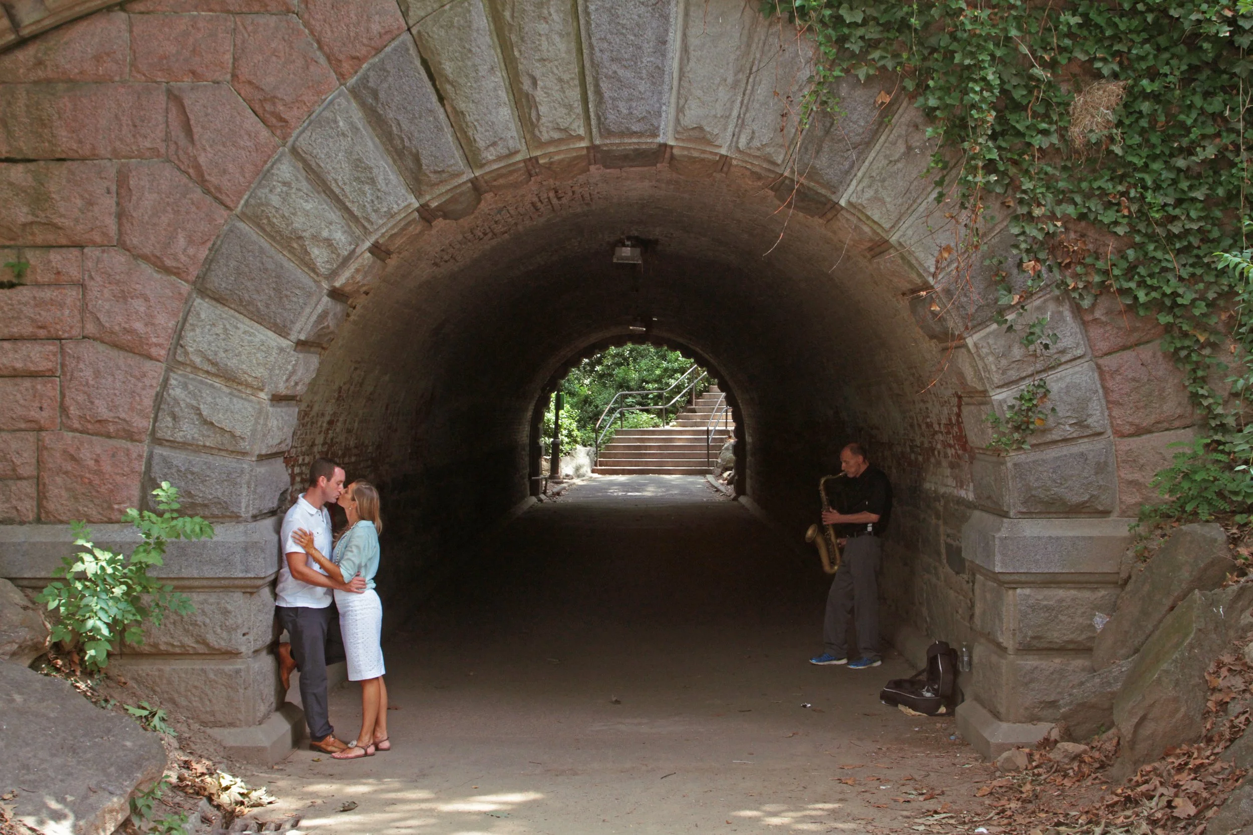 A couple is standing close together and kissing under a stone arch tunnel, while a man playing a saxophone is positioned nearby outside the tunnel.