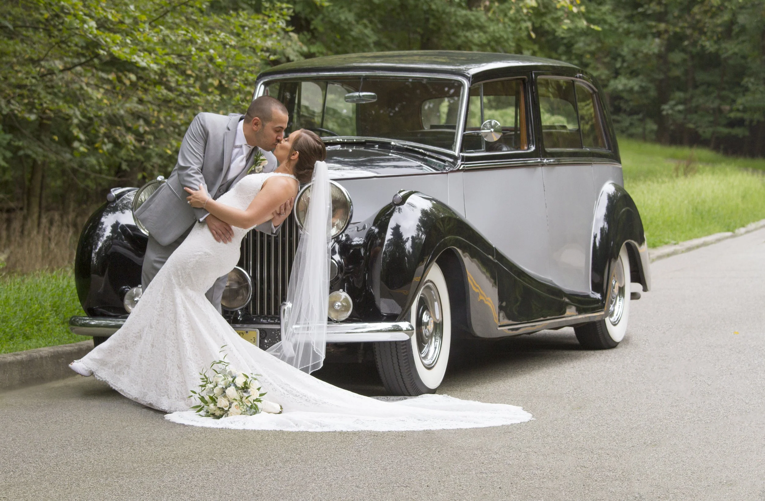 A newlywed couple sharing a kiss in front of a vintage black and gray car, with the bride in a white lace wedding gown holding a bouquet of white flowers, and the groom in a light gray suit, on a roadside with greenery in the background.