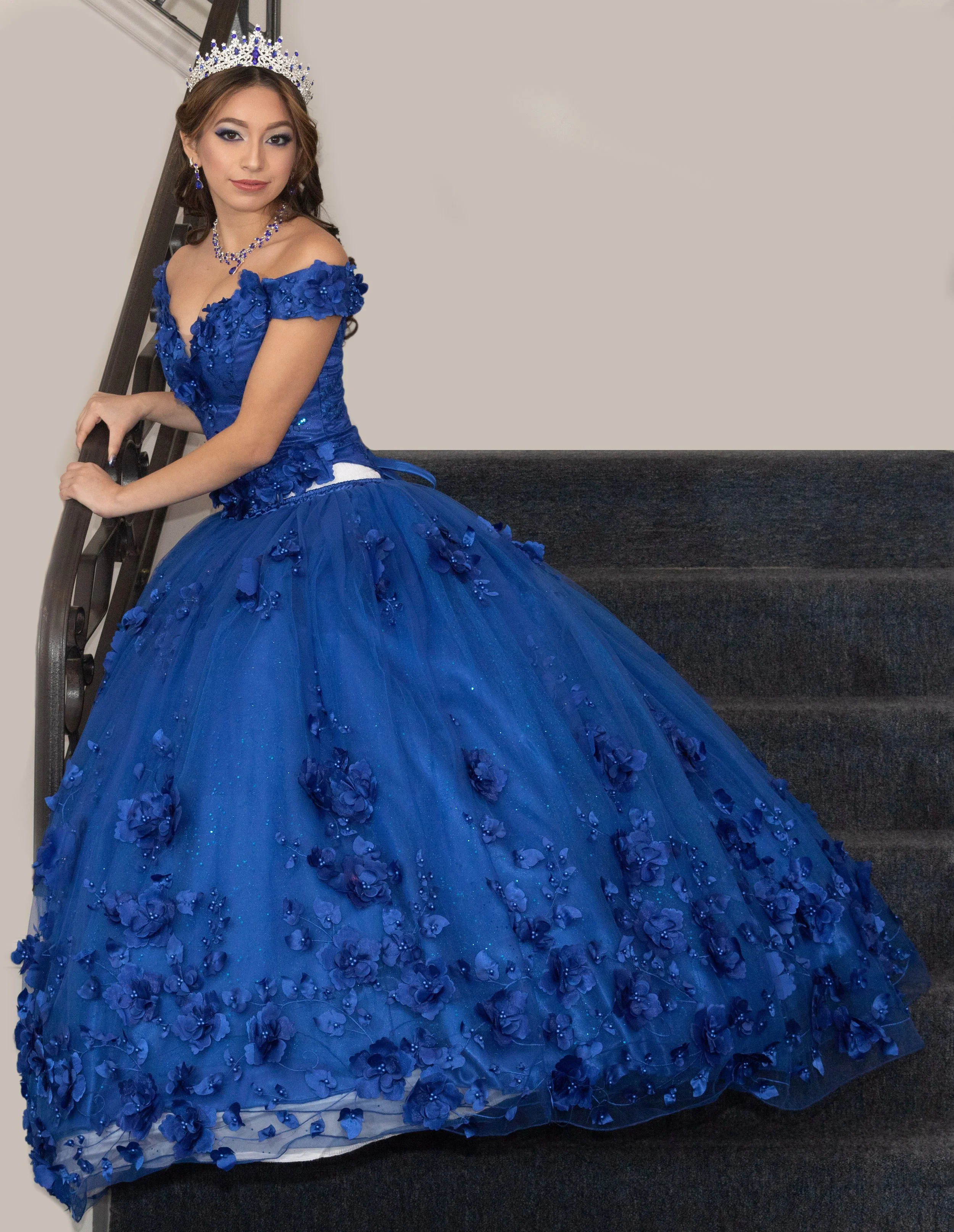 A young quinceañera woman in a regal blue gown adorned with 3D floral embellishments and lace details, standing on a staircase with a black railing, wearing a silver tiara and matching jewelry, posing confidently with her hands on the railing.