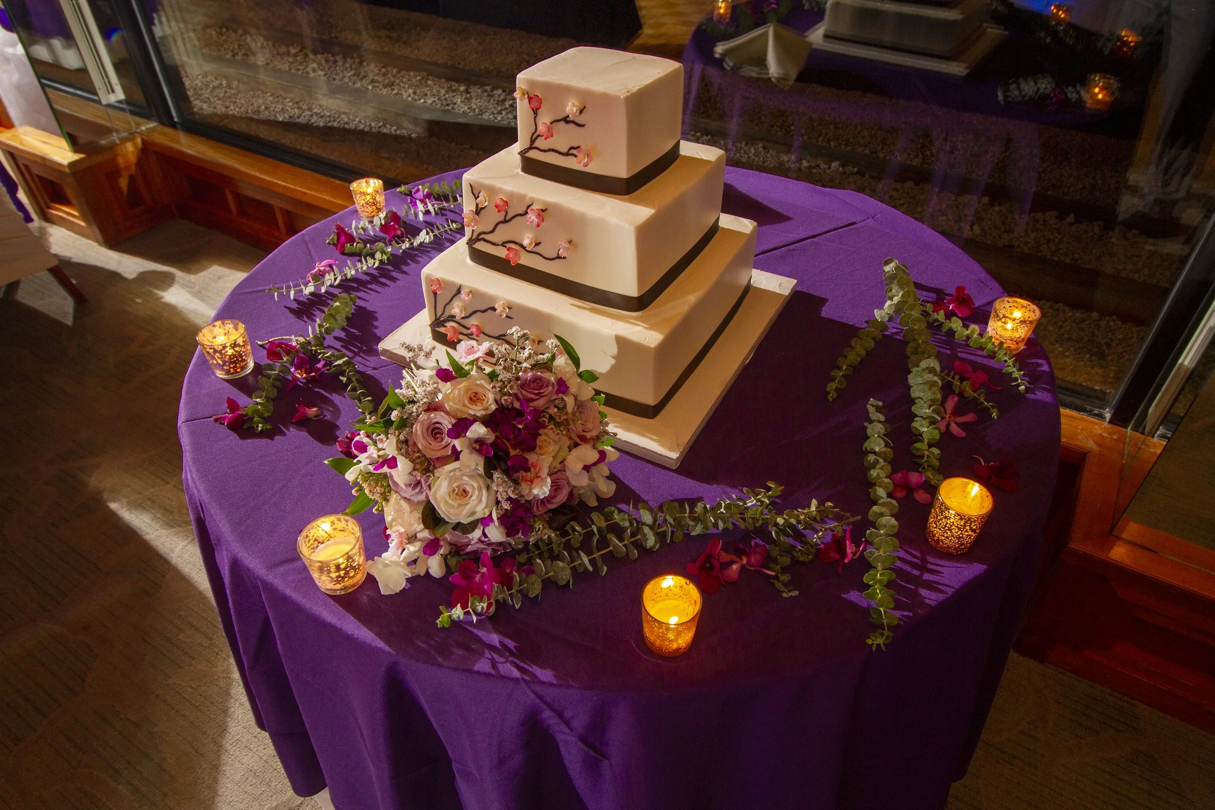 A tiered wedding cake at the Views decorated with pink flowers and branches, placed on a purple tablecloth. The table is surrounded by candle holders with lit candles and a bouquet of pink and white flowers, with greenery scattered on the table.