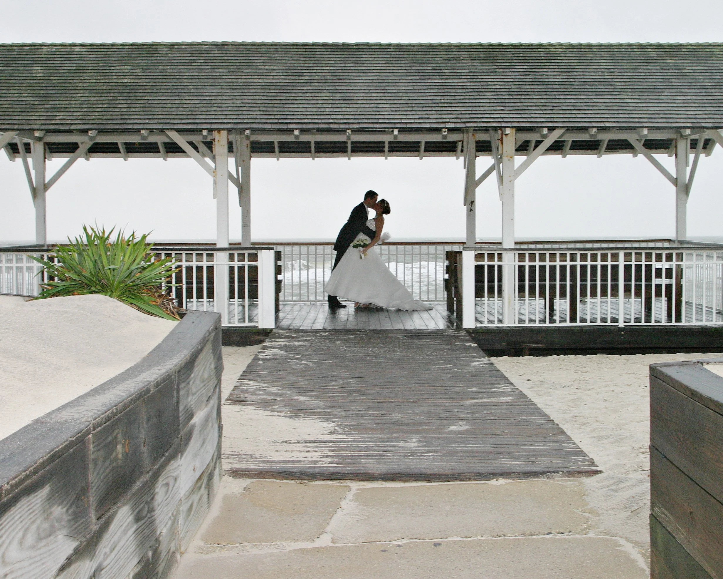 A bride and groom sharing a kiss on a wooden pier or deck at the beach, with the ocean visible in the background, under a pavilion with a sloped roof.