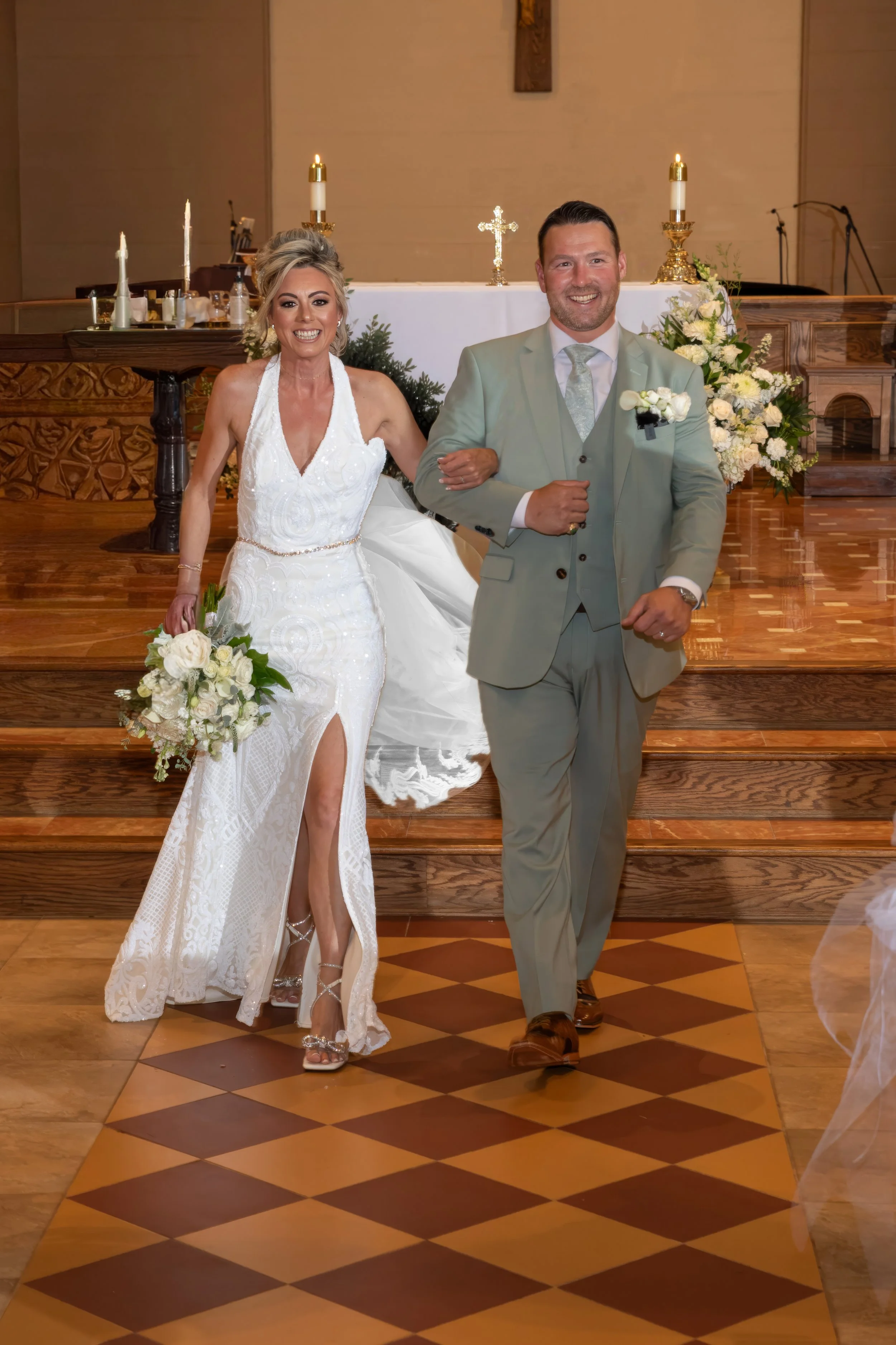 A bride and groom smiling and holding hands during their wedding ceremony inside a church, with an altar decorated with flowers and candles in the background.
