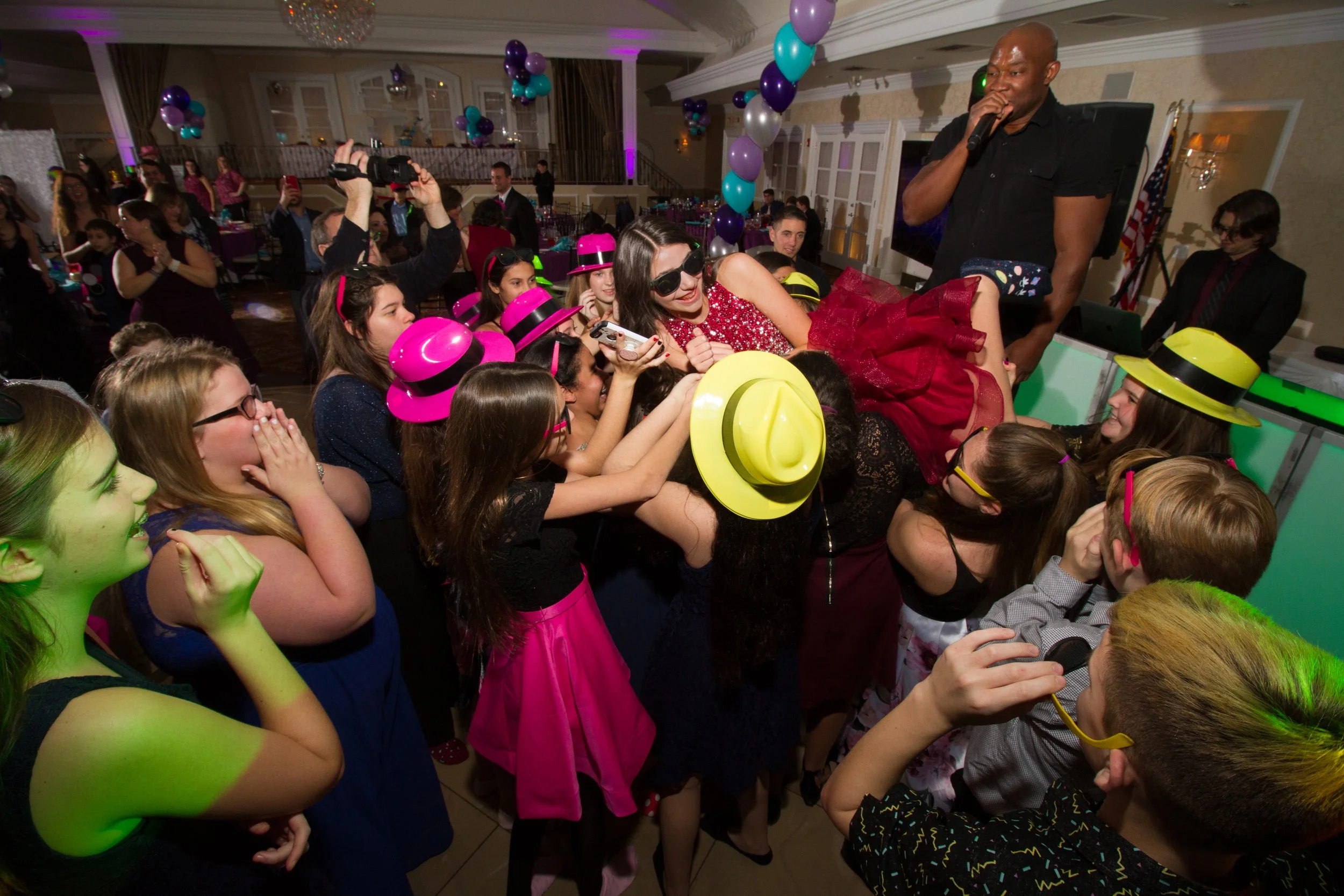 Group of children lifting woman on a chair at a Bat Mitzvah party, wearing sunglasses and colorful hats, with a DJ and balloons in the background.