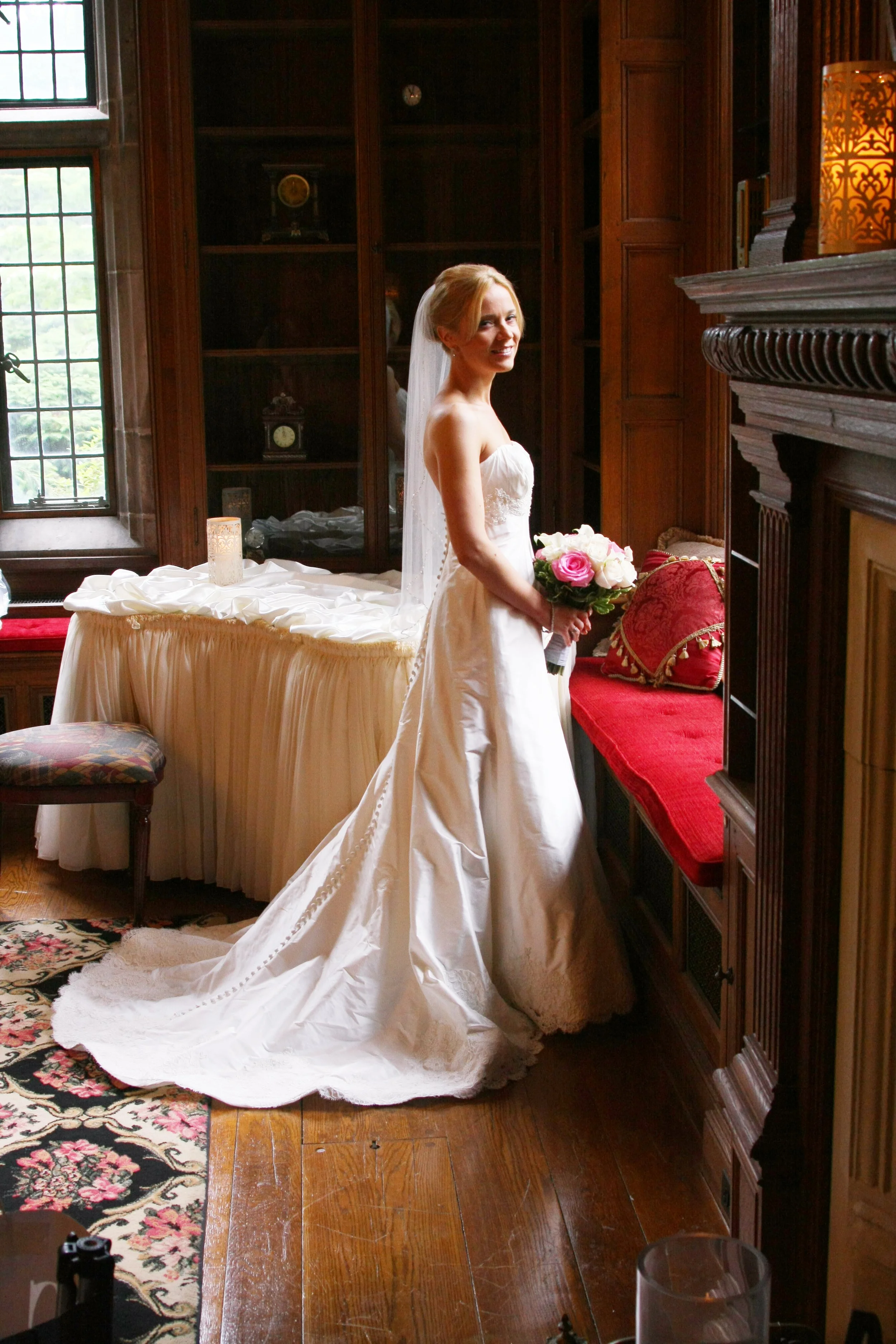 A bride in a white wedding dress holding a bouquet of pink and white roses, standing by a window in a wood-paneled room with a fireplace and a red cushioned window seat.