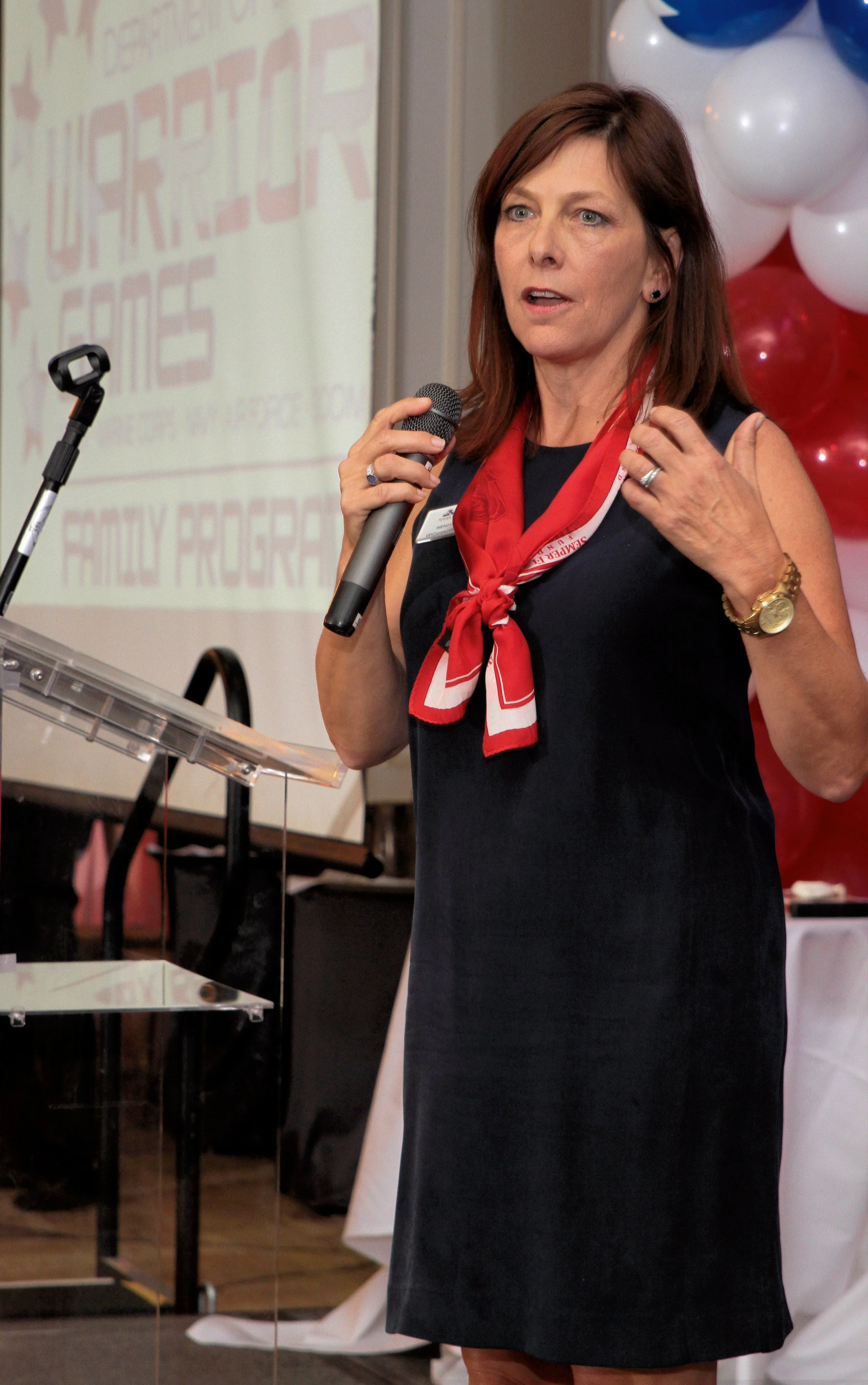 A woman in a black dress speaking into a microphone during a formal event with balloons in the background.