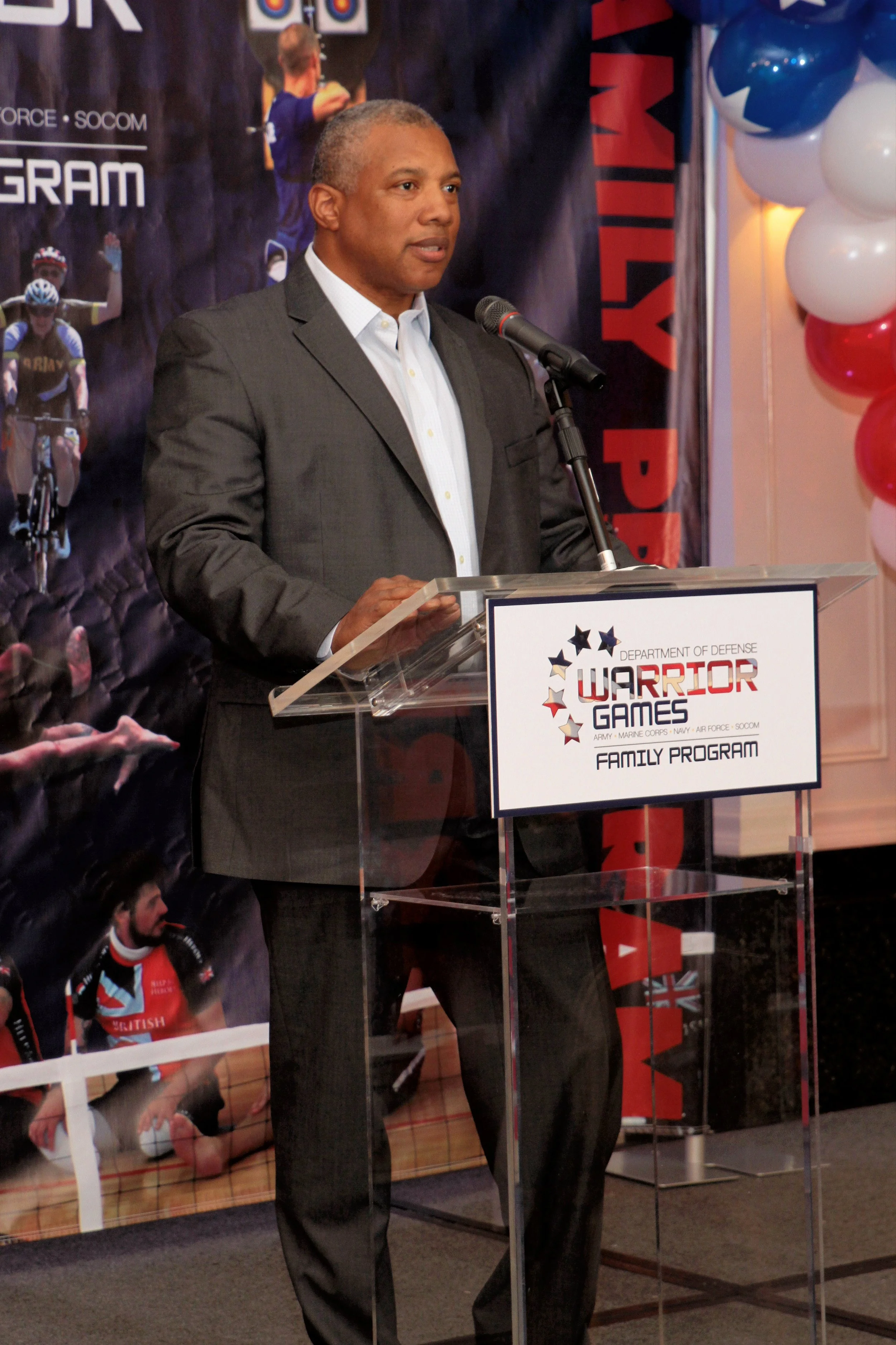 A man in a suit giving a speech at a podium during an event related to the Warrior Games, with balloons and a poster in the background.