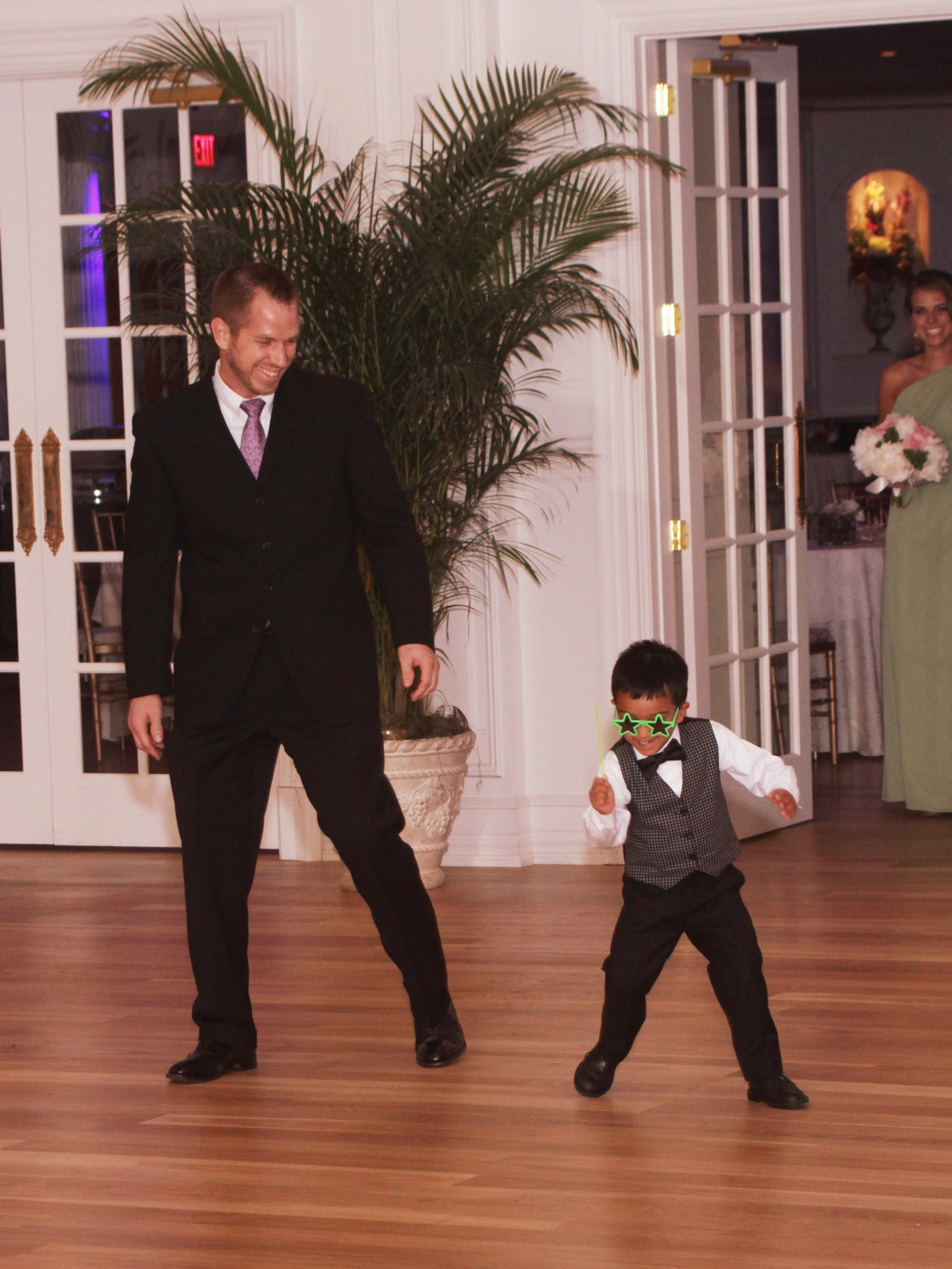 A man in a black suit and a young boy in a black vest and bow tie dancing together, both smiling, in a decorated indoor space with wooden floors, a large potted plant, and women in the background.