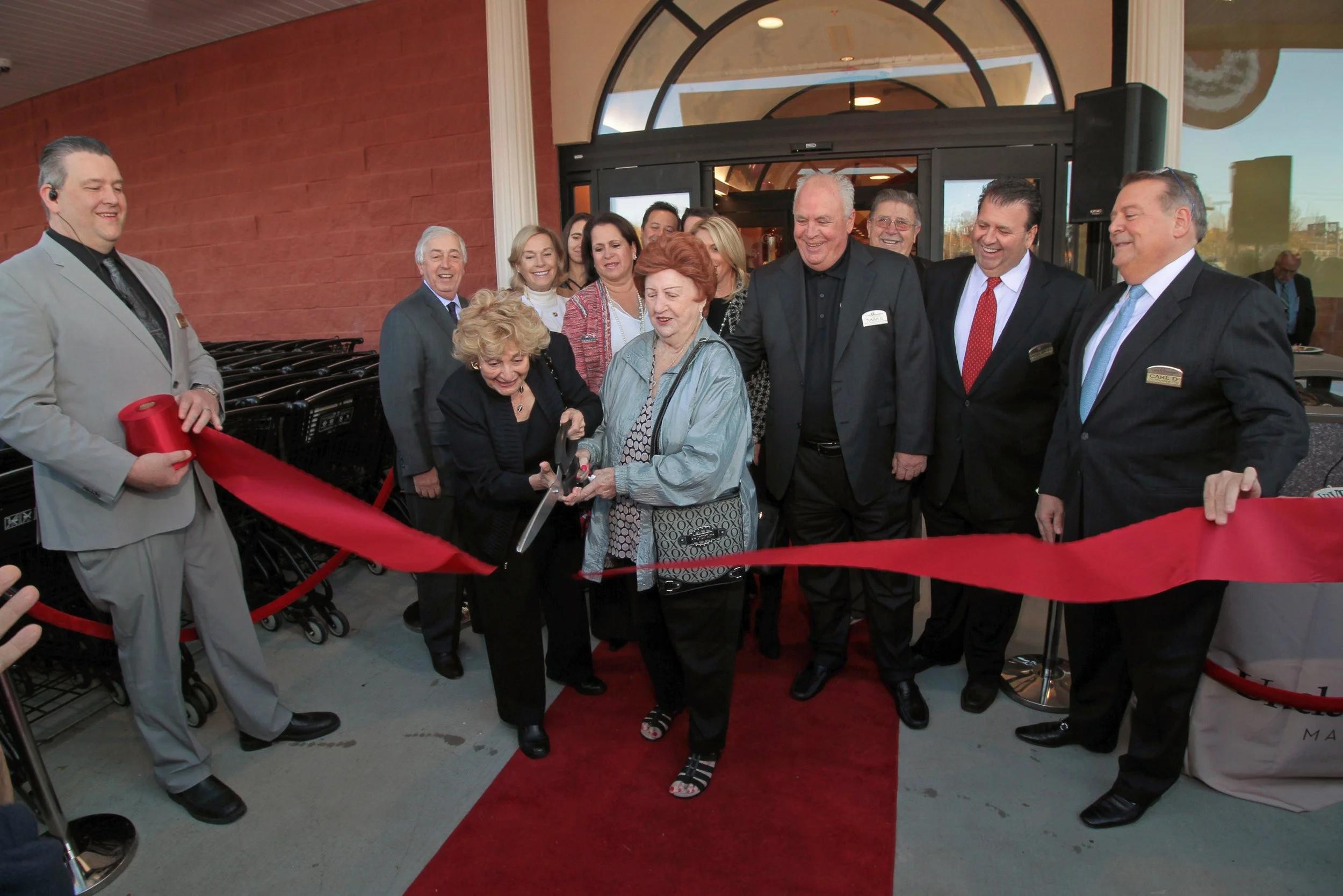 Group of people at a ribbon-cutting ceremony outside a building, with two women in the center holding scissors, surrounded by smiling men and women dressed in formal attire.