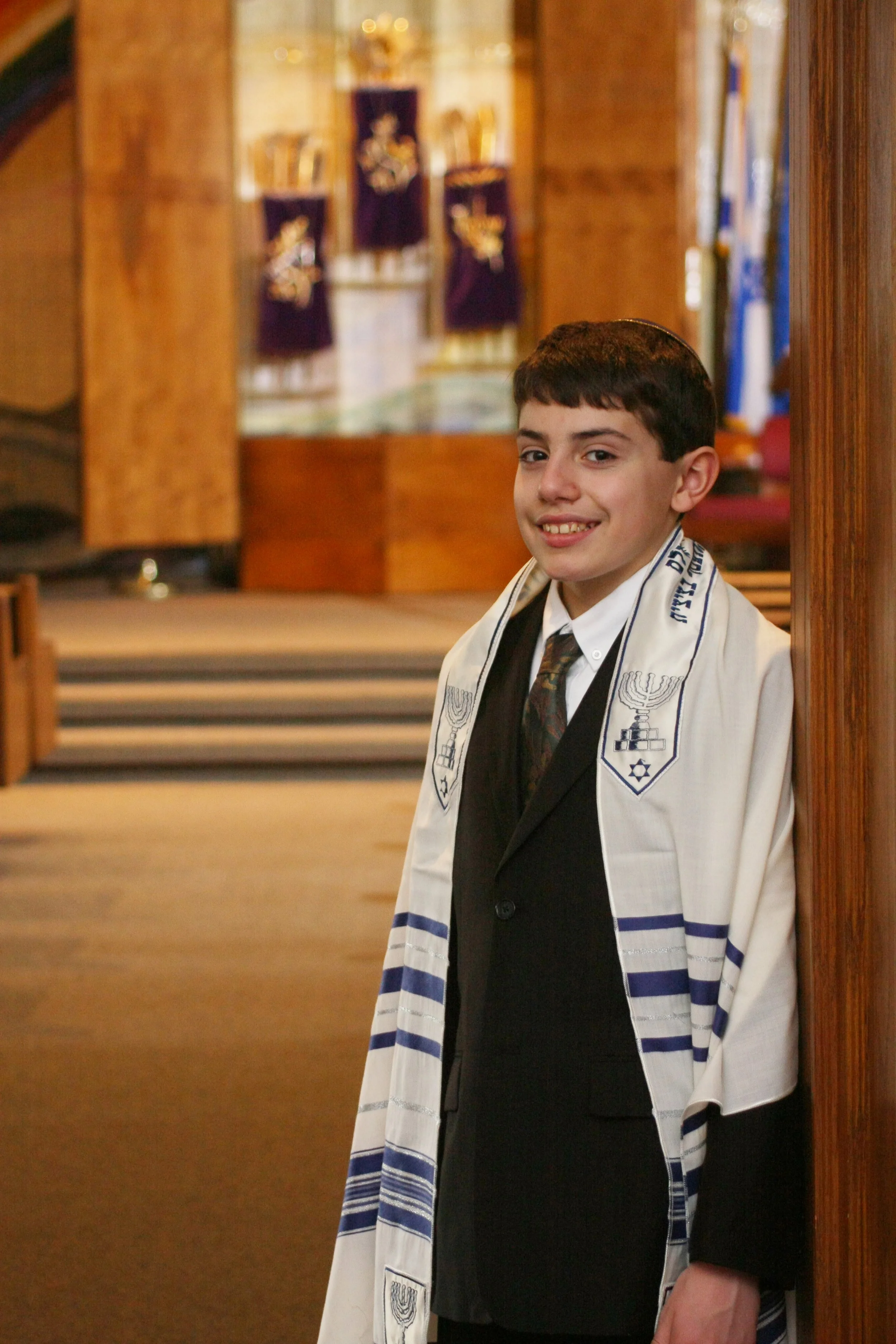 A young boy dressed in formal attire standing inside at Barnert Temple in Franklin Lakes NJ, wearing a tallit (Jewish prayer shawl) over his shoulders, with a slight smile and leaning against a wooden wall.