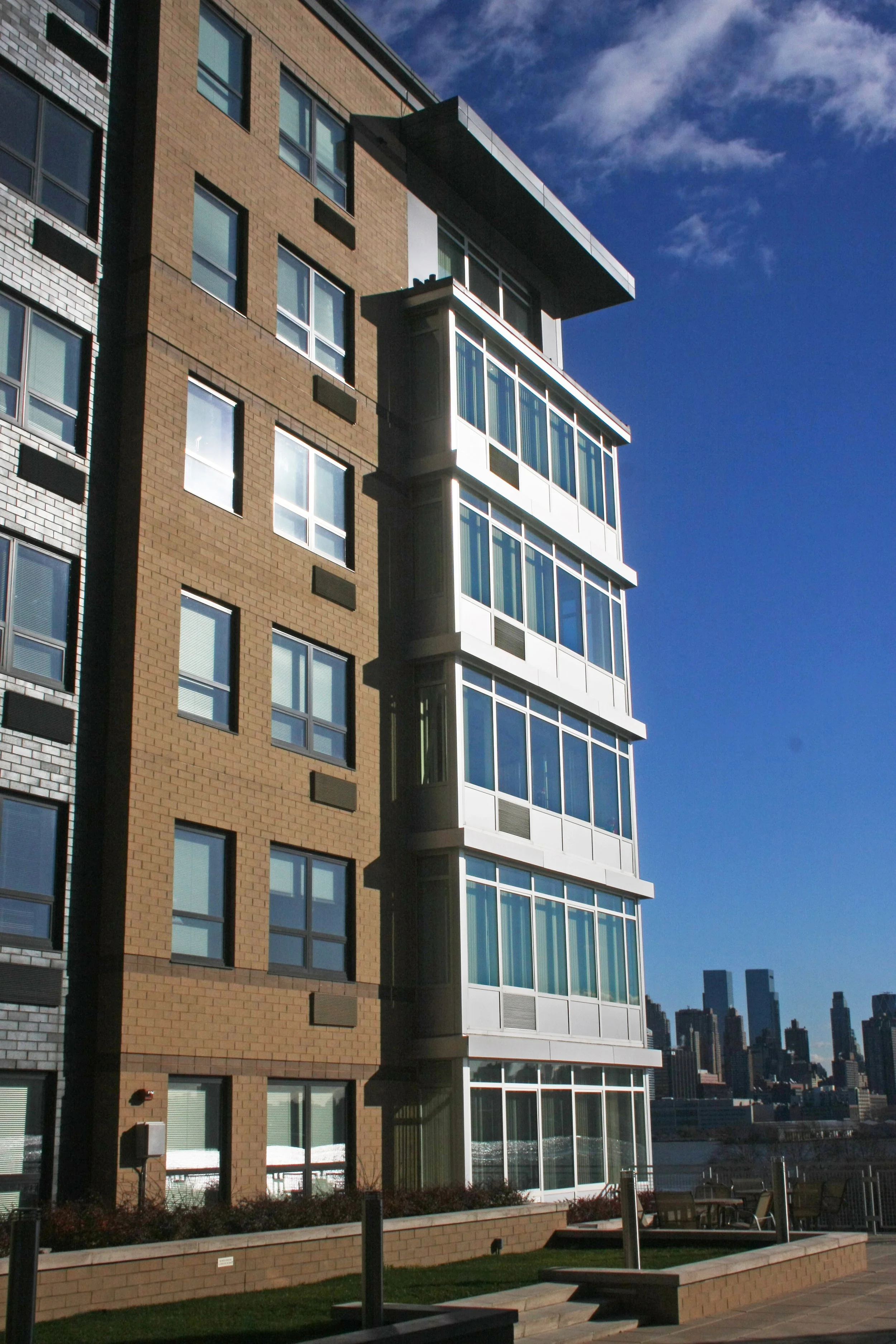 A modern multi-story apartment building with a brown brick facade and a glass-enclosed balcony section, set against a clear blue sky with a distant city skyline.