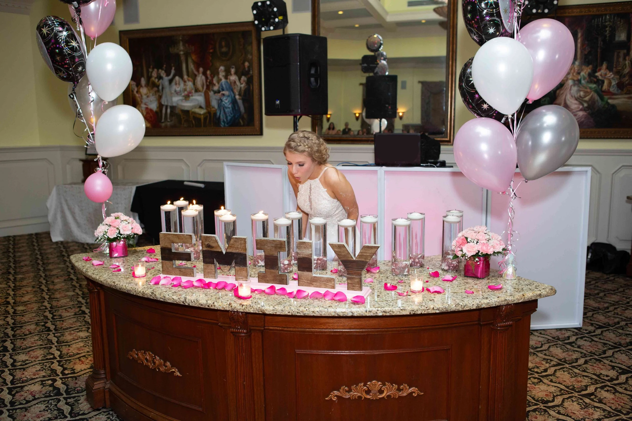 A woman in a white dress at the Brick house of Wyckoff leans over a decorated table at a celebration, with balloons, candles, flowers, and a wooden 'EMILY' sign on the table. A large mirror and paintings are seen in the background.