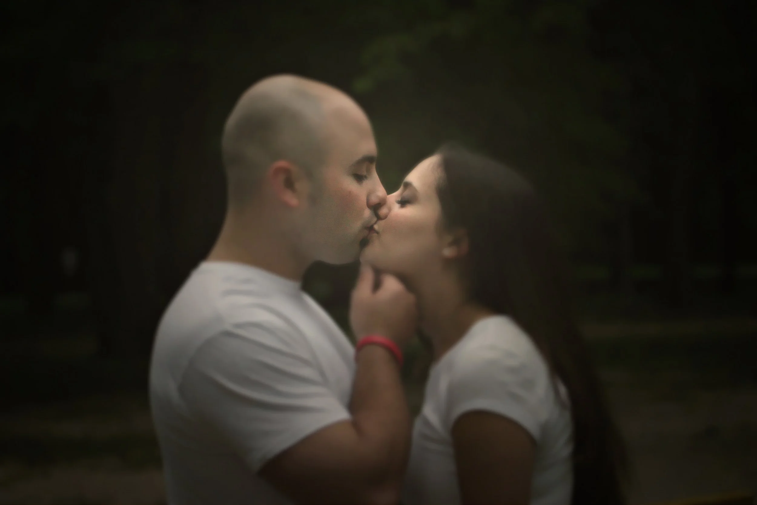A man and woman sharing a kiss outdoors at night, with dark trees in the background. They are both wearing white shirts.