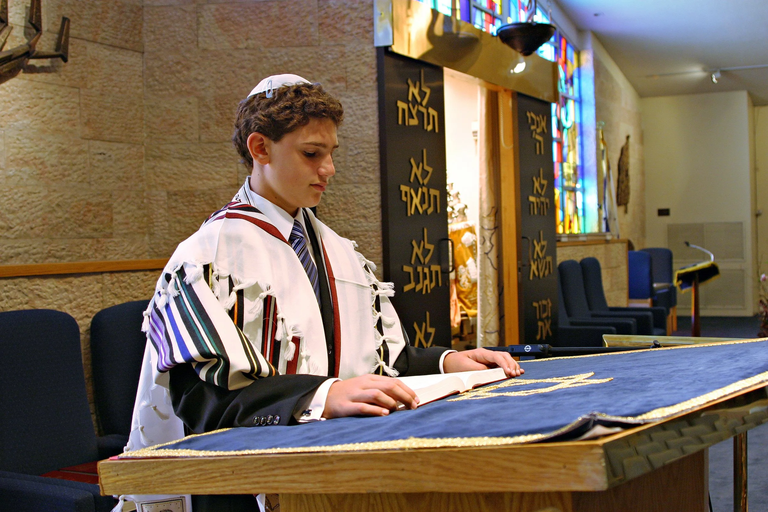 A Bar Mitzvah boy wearing Jewish prayer garments, including a tallit and kippah, reading from a prayer book on the bimah at a synagogue Bar Mitzvah ceremony.