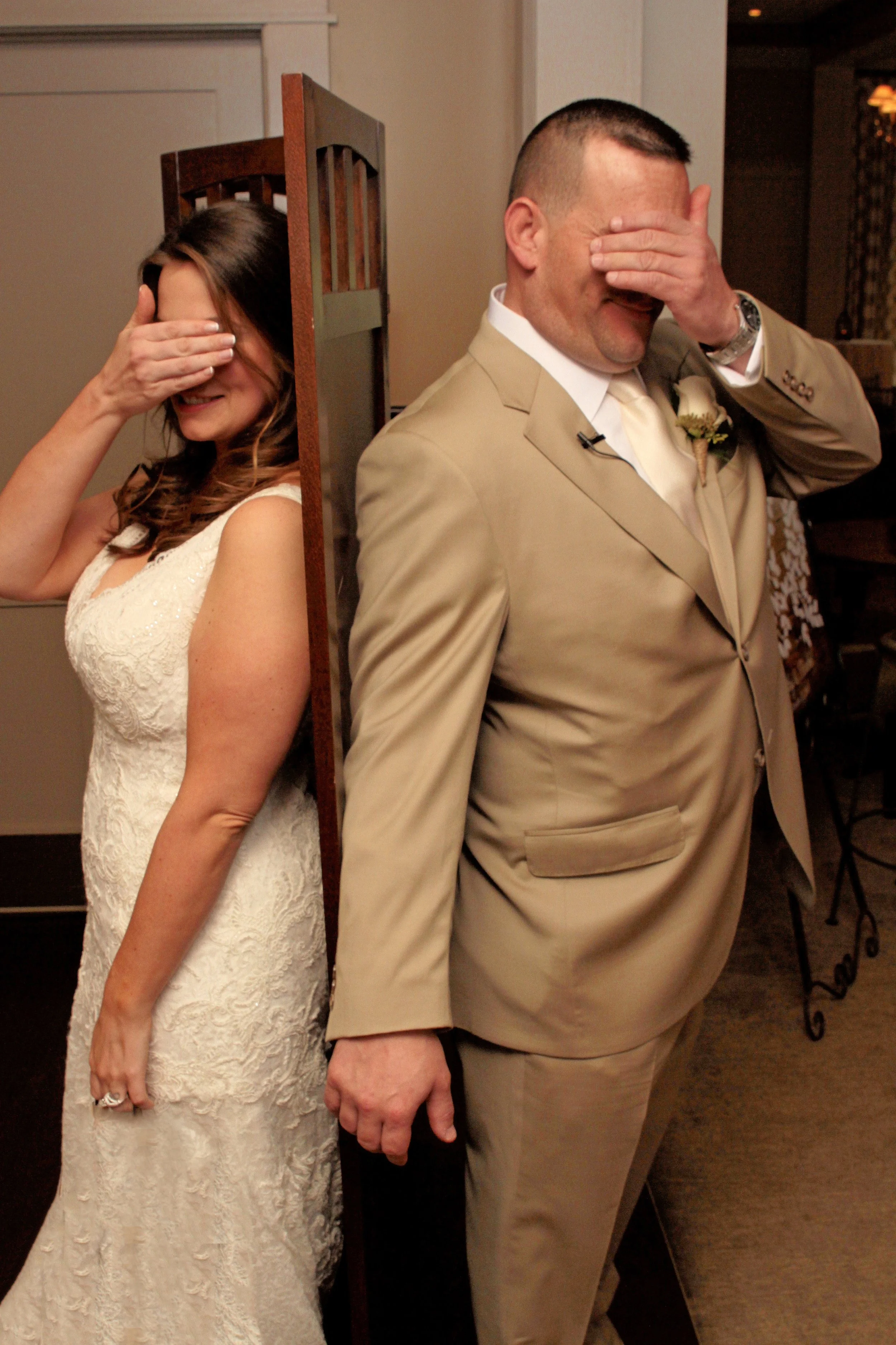 A bride and groom stand back-to-back with a folding screen between them, both covering their eyes and smiling during a wedding game.