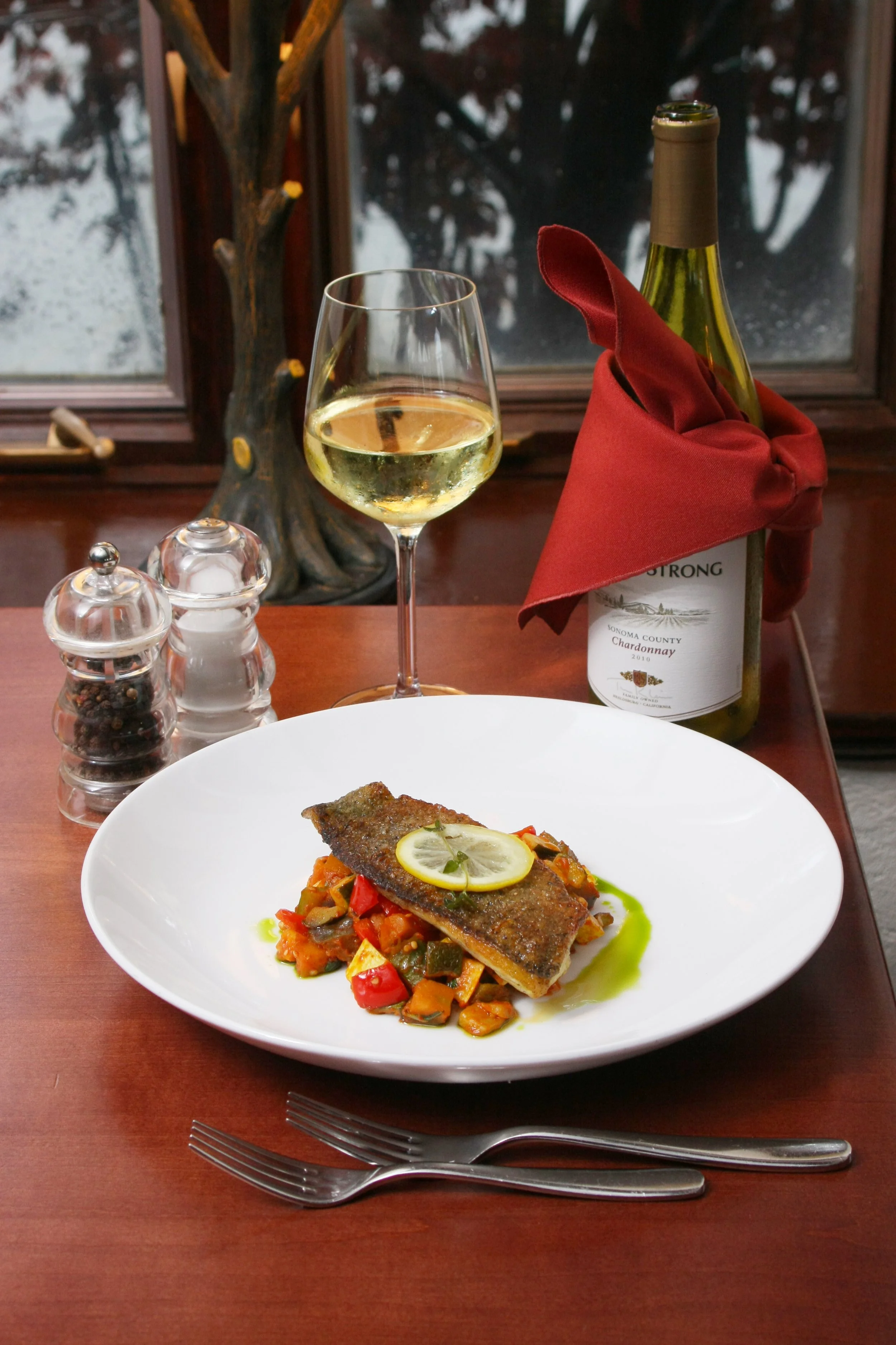 A plated serving of cooked fish topped with a lemon slice, served on a bed of mixed vegetables, with a glass of white wine, a bottle of wine with a red cloth, and salt and pepper shakers on a wooden table near a window.