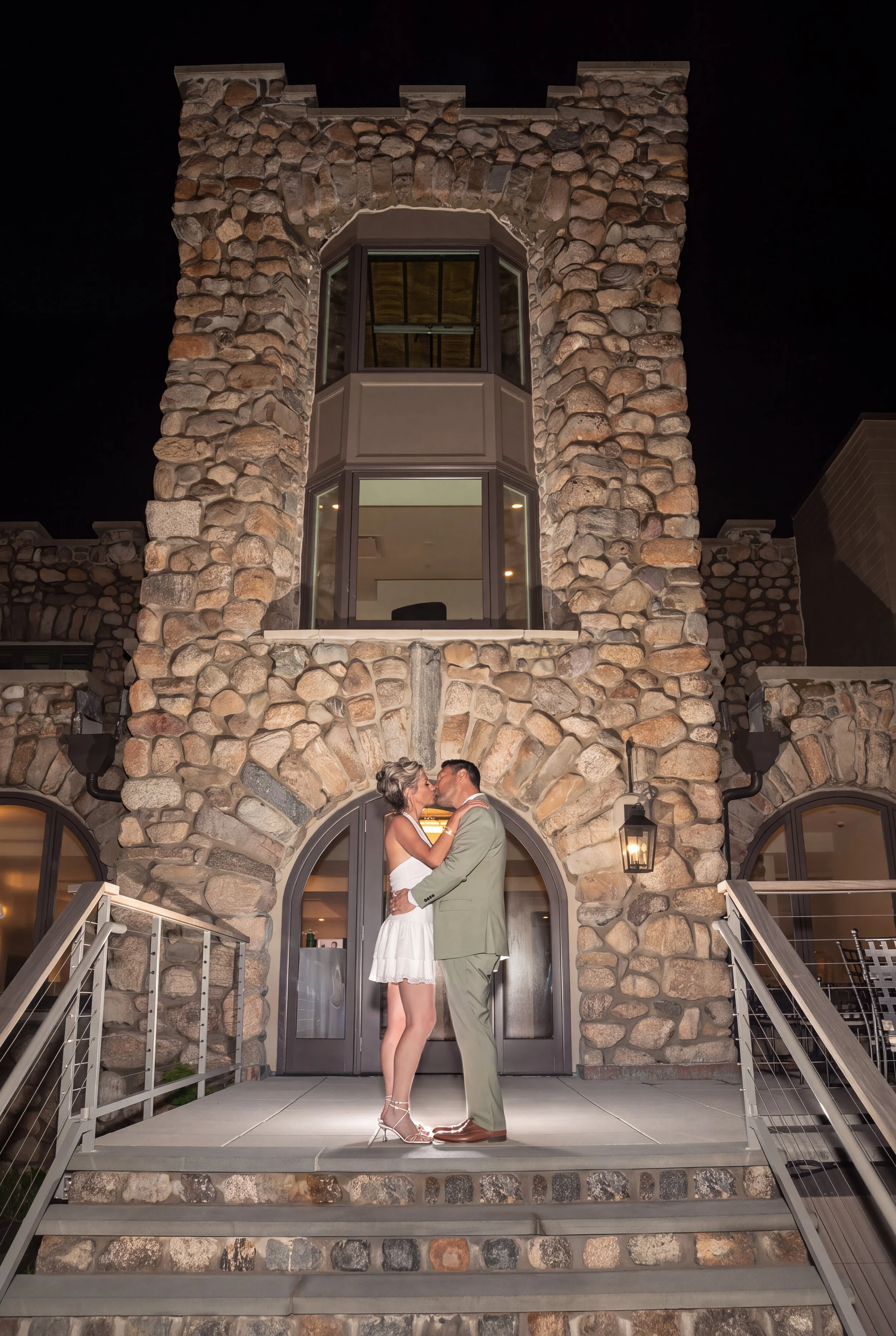 A couple dressed in formal attire kissing on a stone staircase in front of a stone building at night.