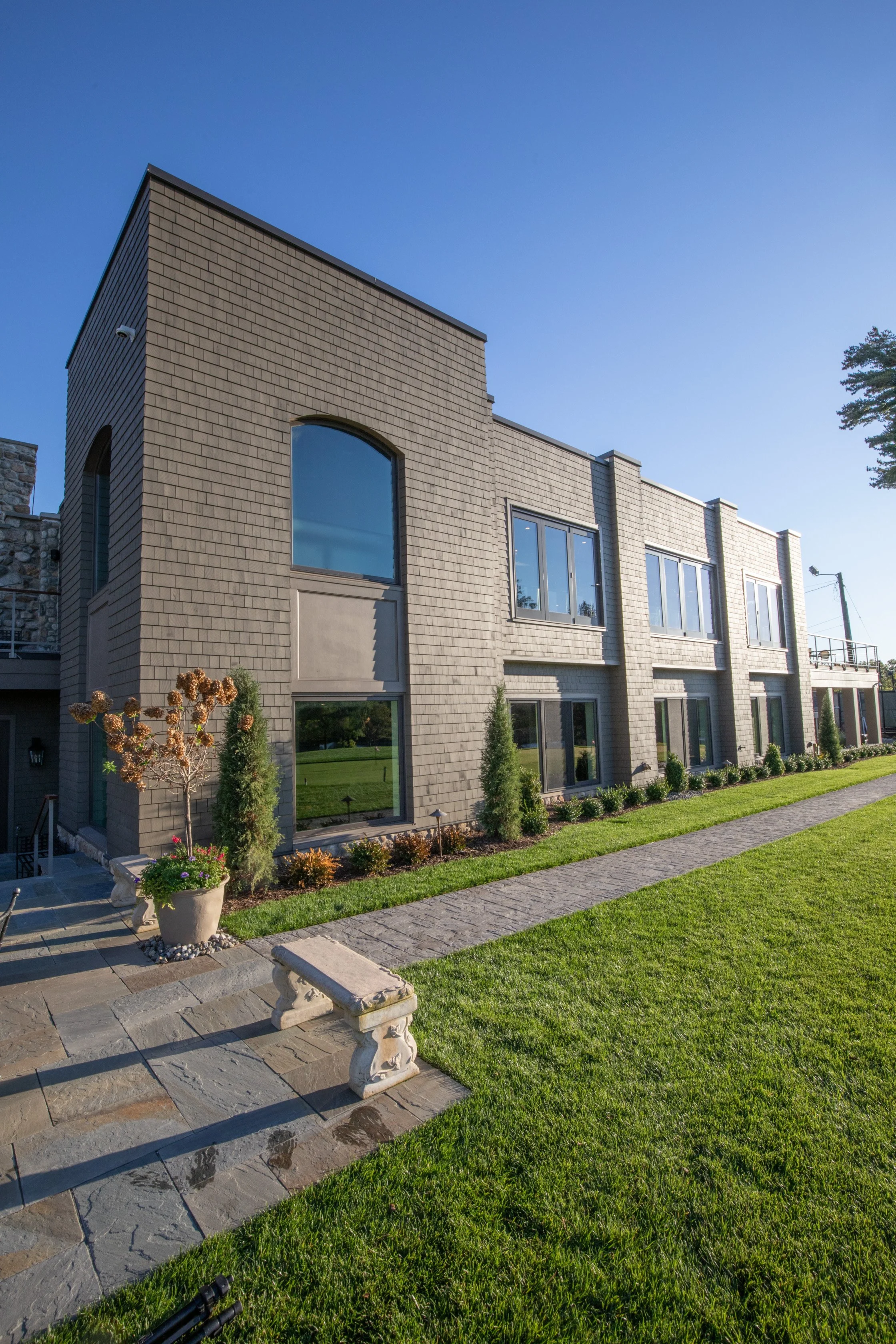 Modern two-story building at the Ramsey Golf and Country Club with gray brick exterior, multiple large windows, and a landscape with green grass, small trees, and shrubs, under a clear blue sky.