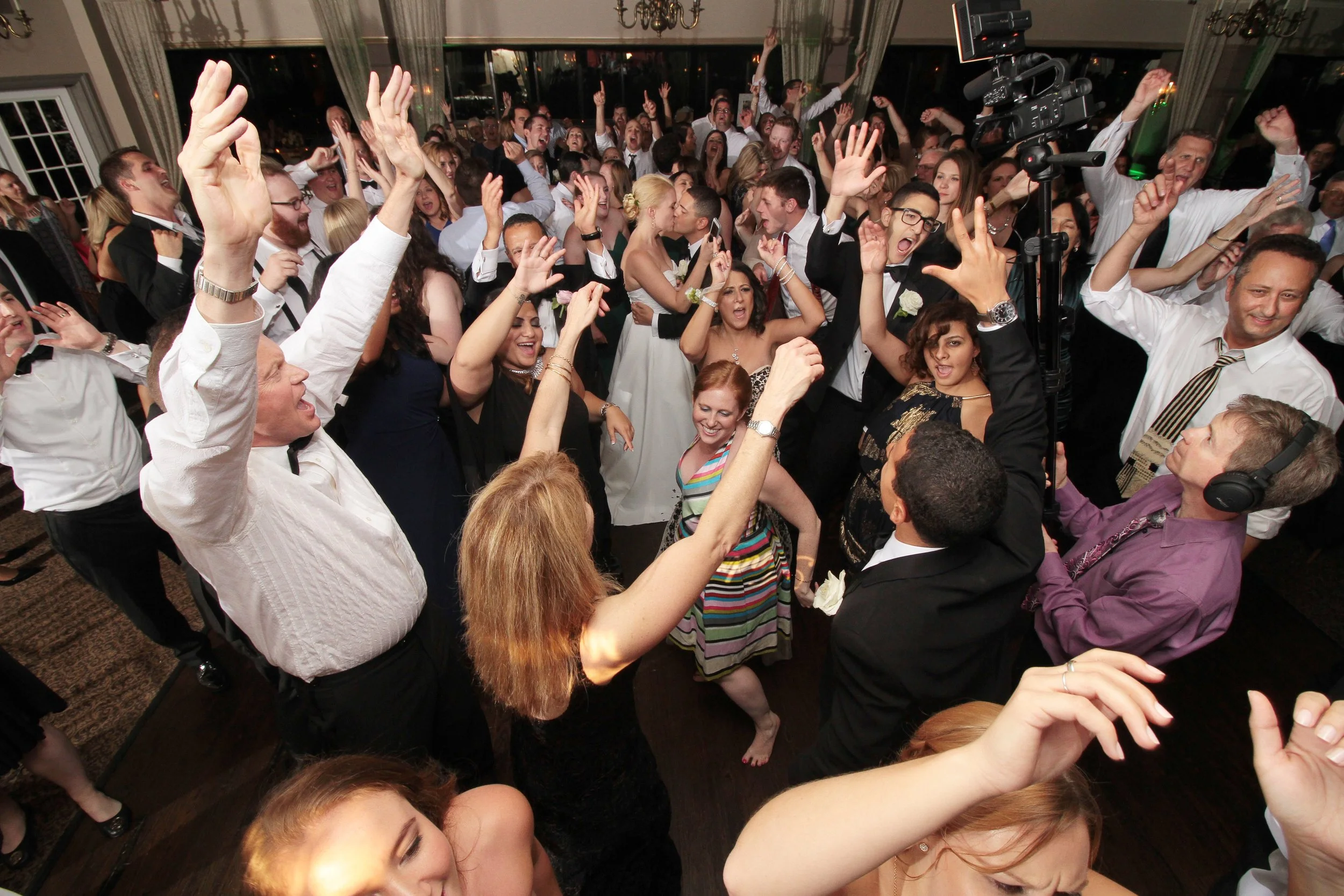 Dance floor with wedding guests dancing, celebrating, and enjoying. Many guests with hands in the air, smiling, and singing along in a lively indoor setting.