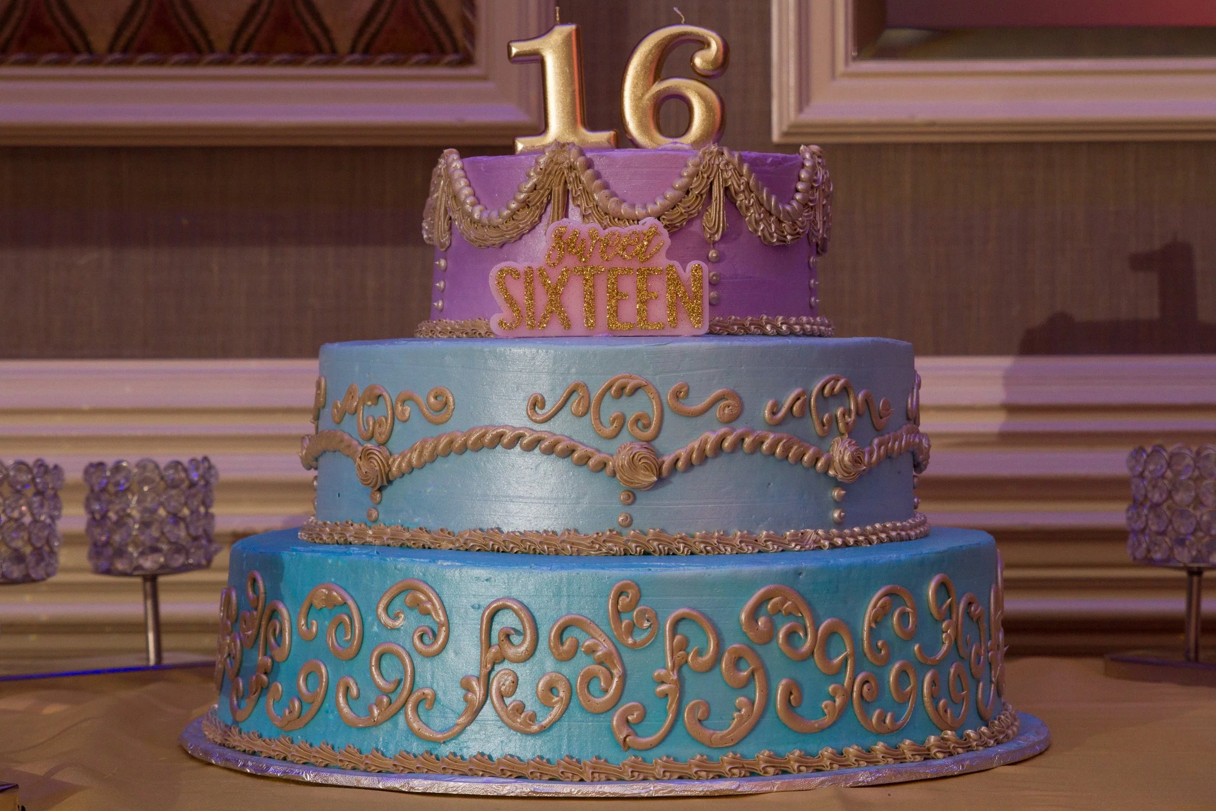 Three-tiered sweet 16 birthday cake with blue and purple frosting, adorned with gold and pink decorations, topped with a gold "16" candle, and says "Sweet Sixteen".