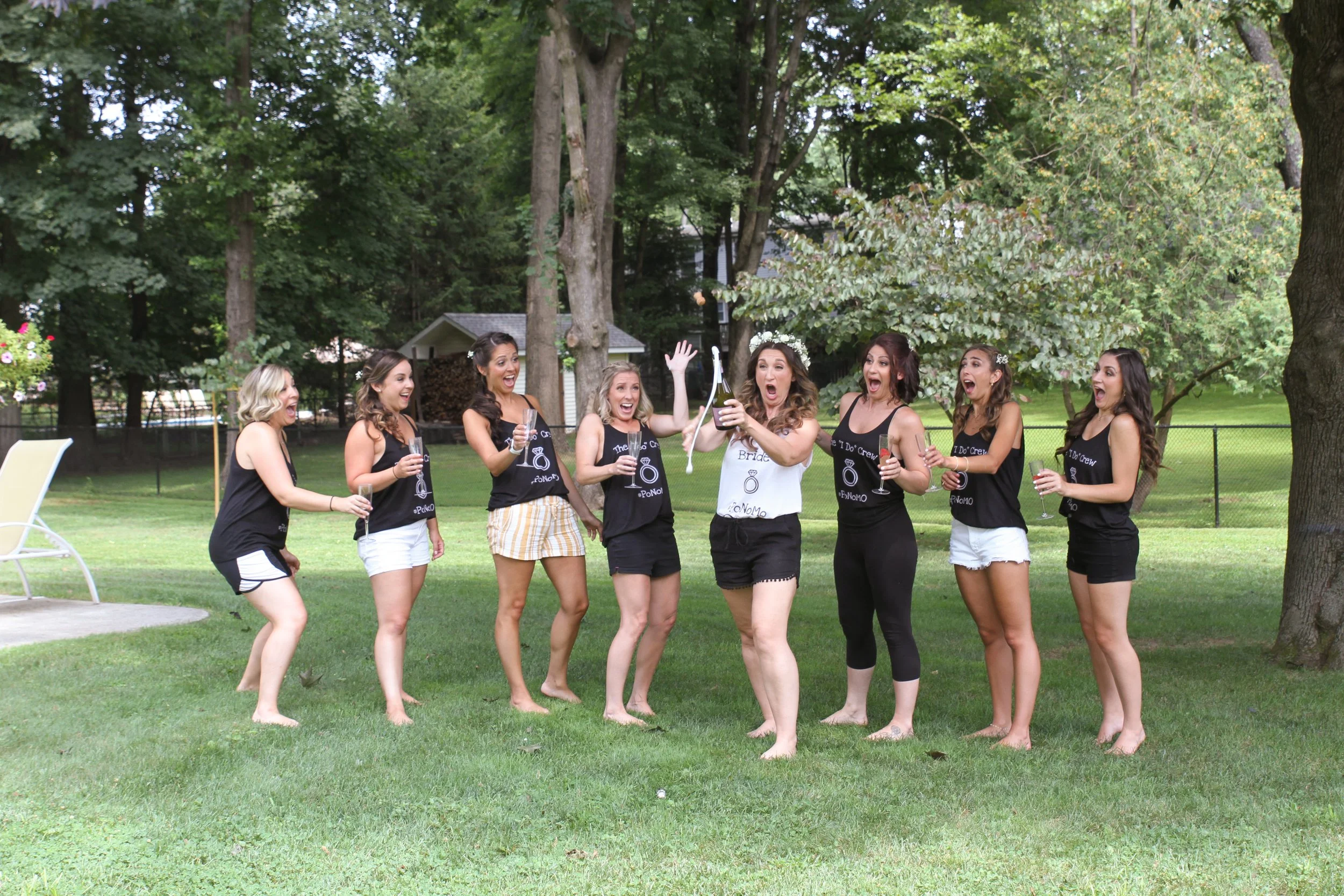 A group of women celebrating outdoors; one woman opens a champagne bottle, all appear happy and excited, standing on grass with trees and a small building in the background.