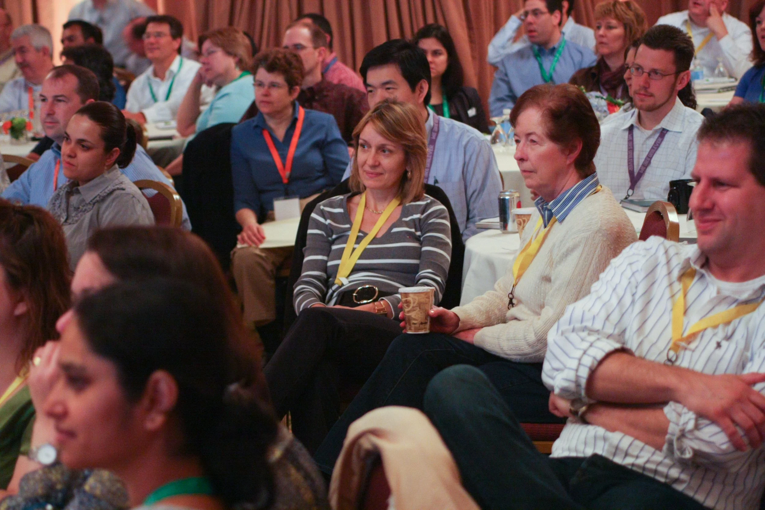 Audience at a conference or seminar listening attentively, sitting at round tables with some holding coffee cups and cans, in a dimly lit room.