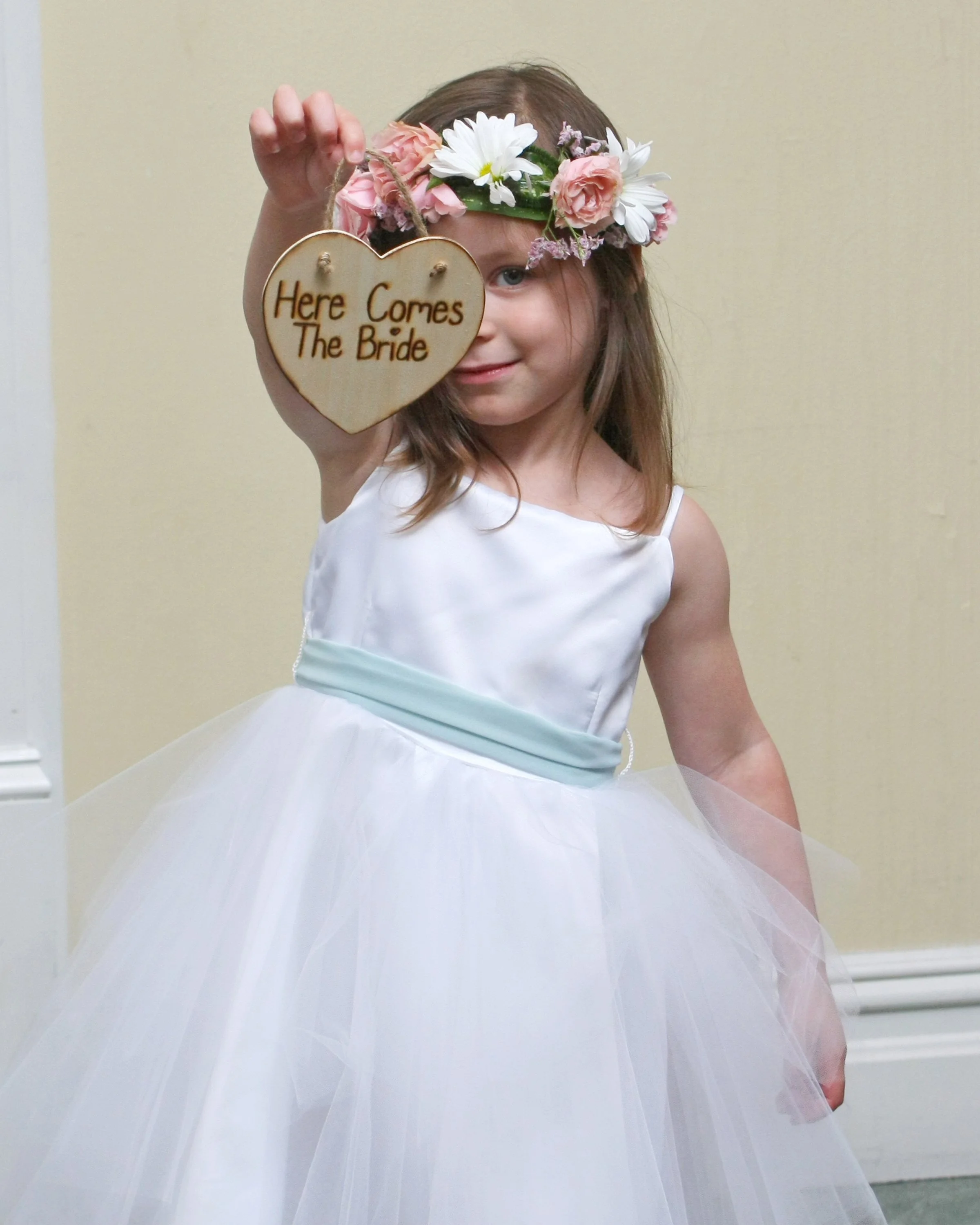A young flowergirl wearing a white dress and floral crown holding a heart-shaped sign that says 'Here Comes The Bride'.