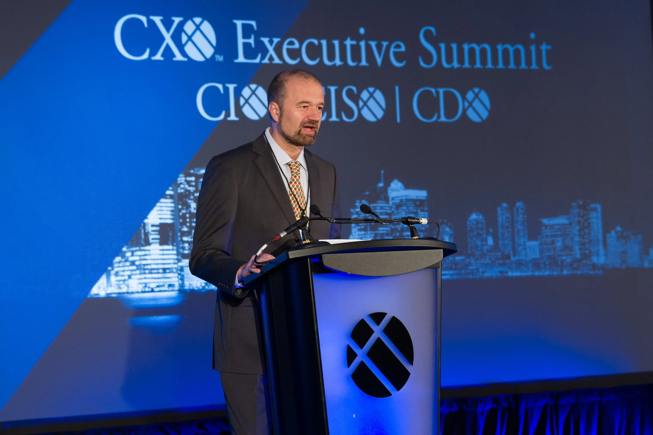 A man in a suit and tie speaking at a podium during the CXO Executive Summit, with a large screen behind him displaying the event's logo and a city skyline.