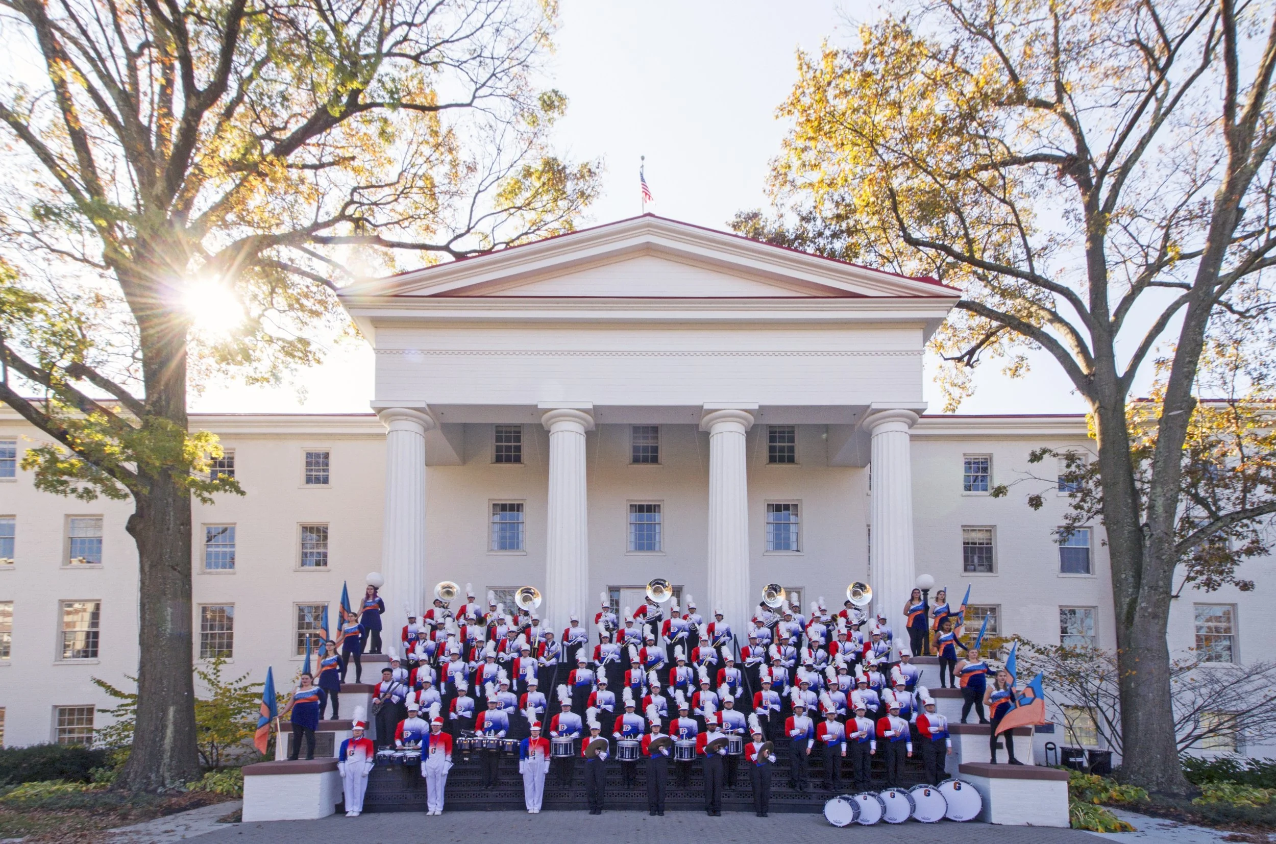 A large marching band performs on the steps of a white building with four columns, during a sunny autumn day with trees around for college website.