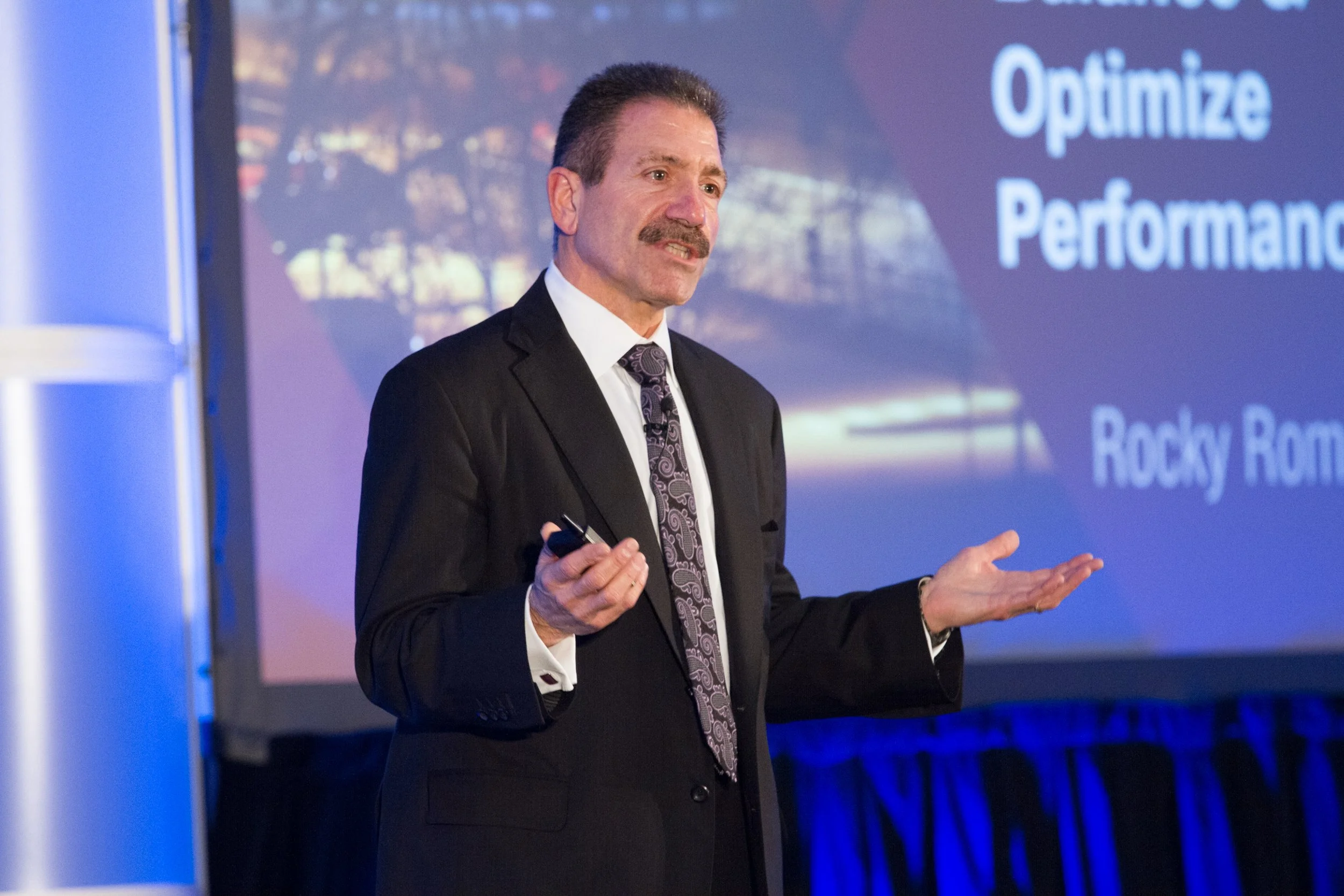 A man in a suit and tie giving a presentation at a business meeting conference in front of a screen displaying the words "Optimize Performance" and "Rocky R". He is gesturing with his hands and holding a remote control.