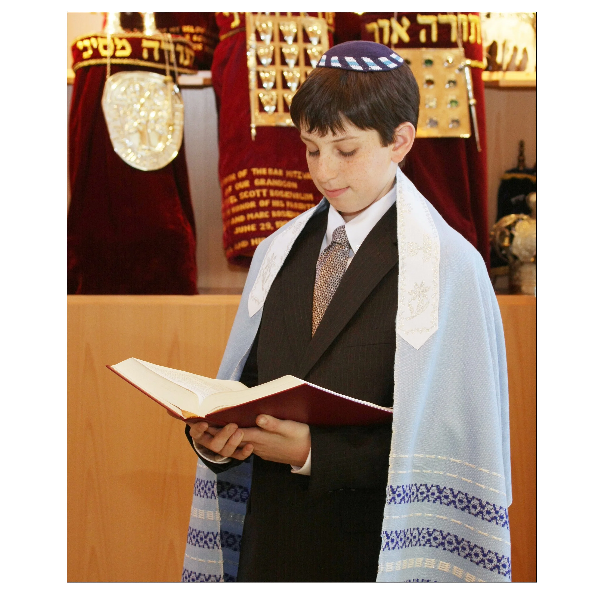 Young Bar Mitzvah boy dressed in a suit and tallit, reading from a prayer book during a ceremony, with religious artifacts and banners in the background.